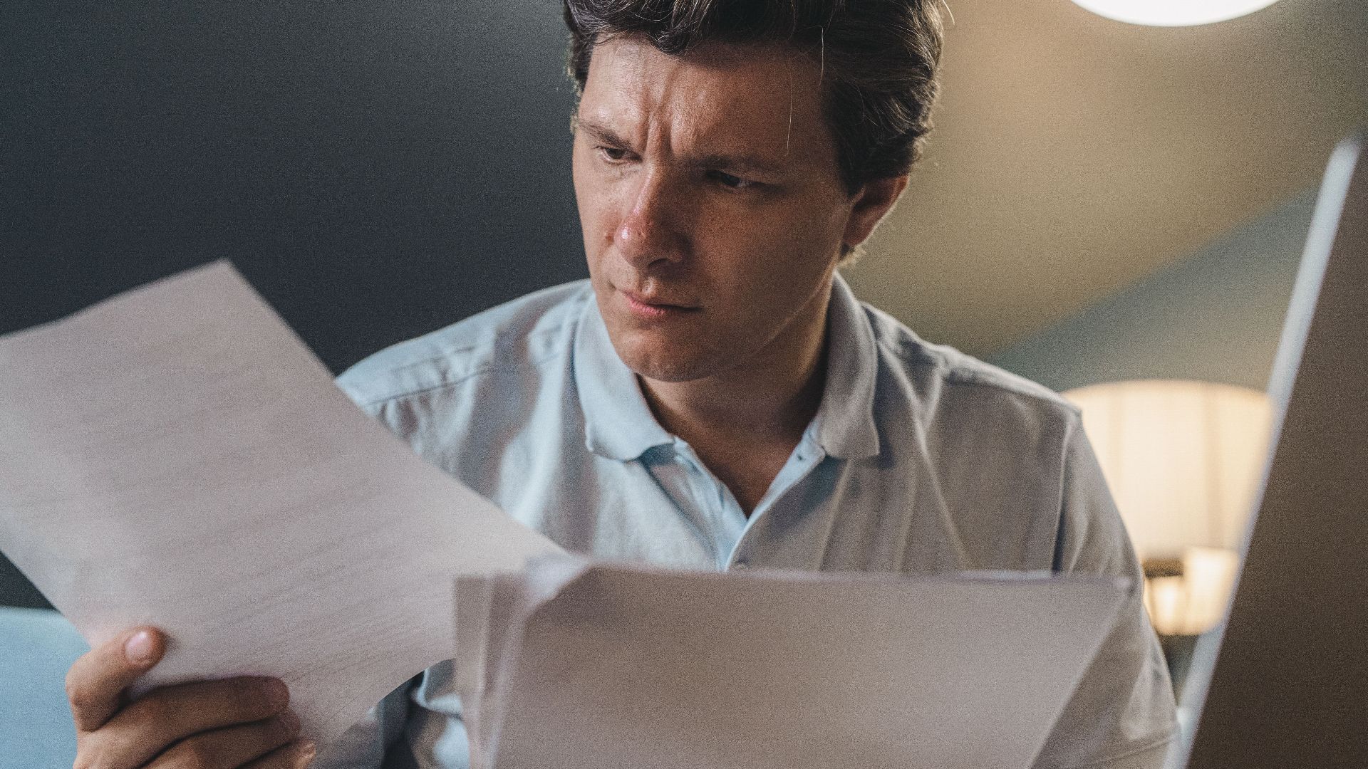 A man intently examines papers, seated indoors under warm lighting, focusing on his work.