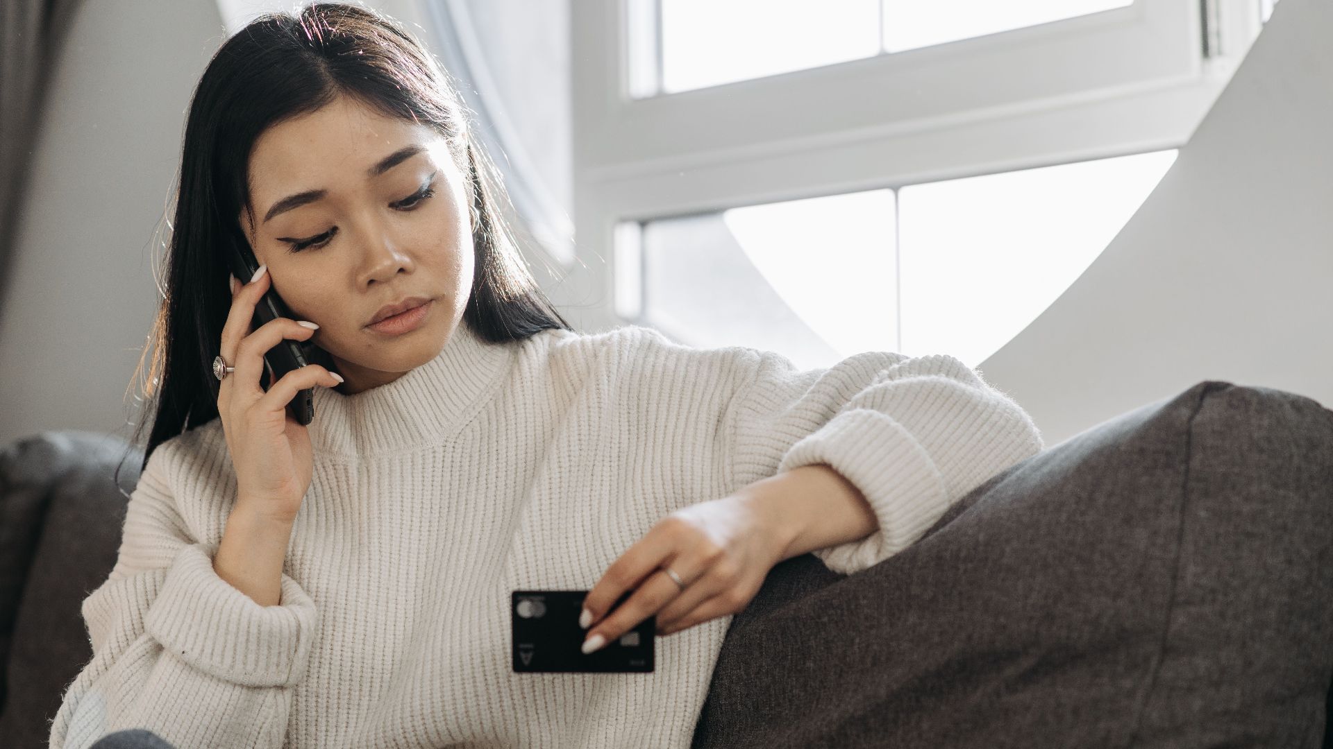 A woman sits on a sofa, multitasking with a phone and credit card, focusing on online shopping.