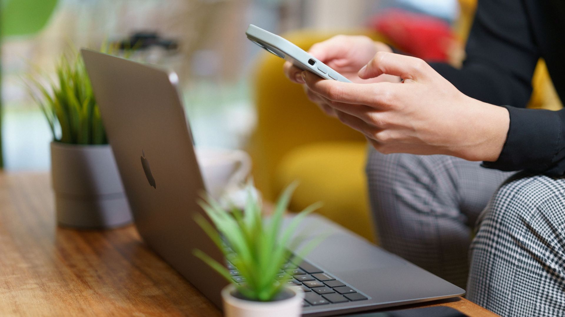 A person working on a smartphone and laptop at home with plants nearby.