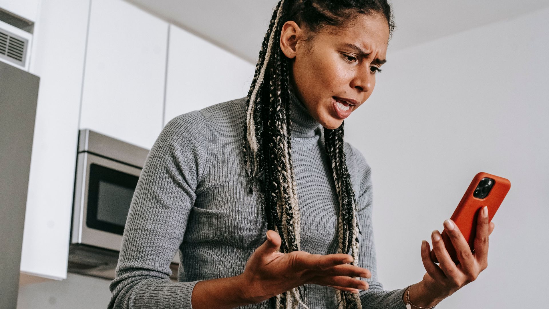 From below of angered black female with Afro braids in casual outfit gesticulating and resenting while having video call on smartphone in modern light kitchen
