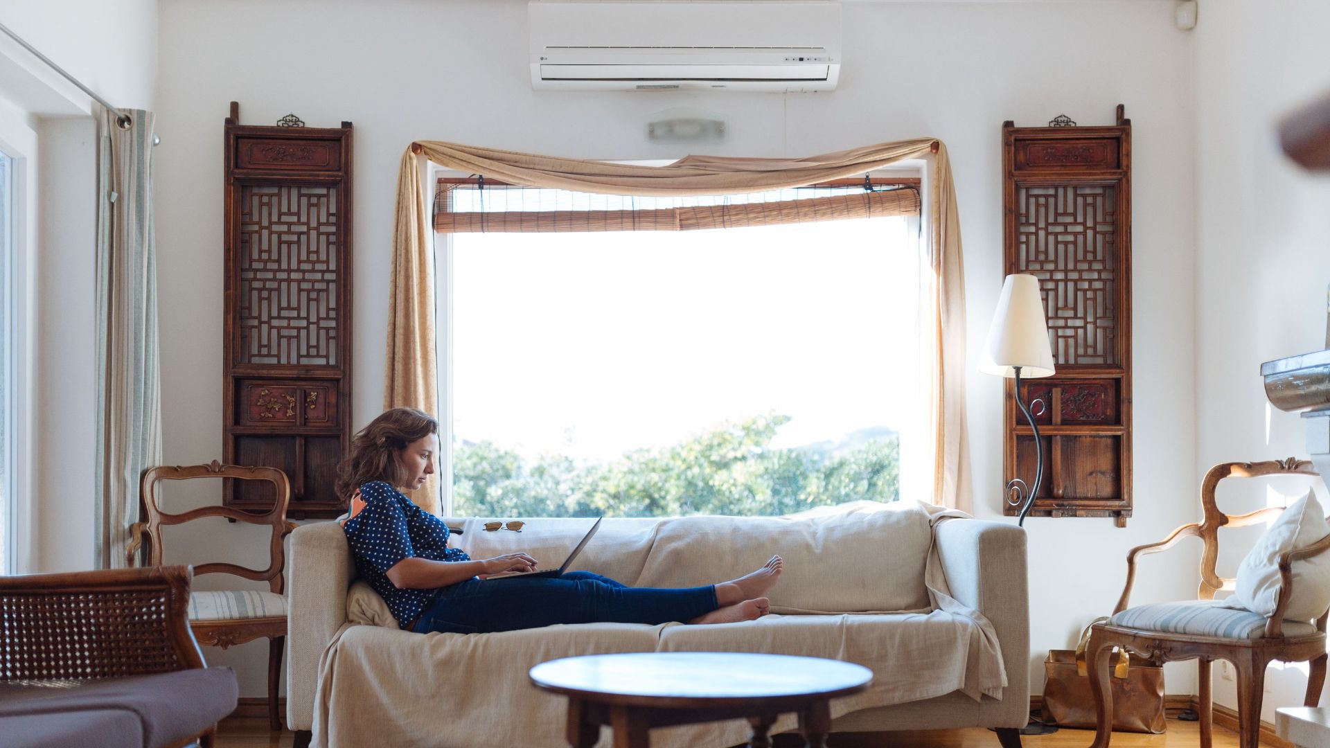 A woman working on a laptop while comfortably lounging on a sofa in a sunlit living room.