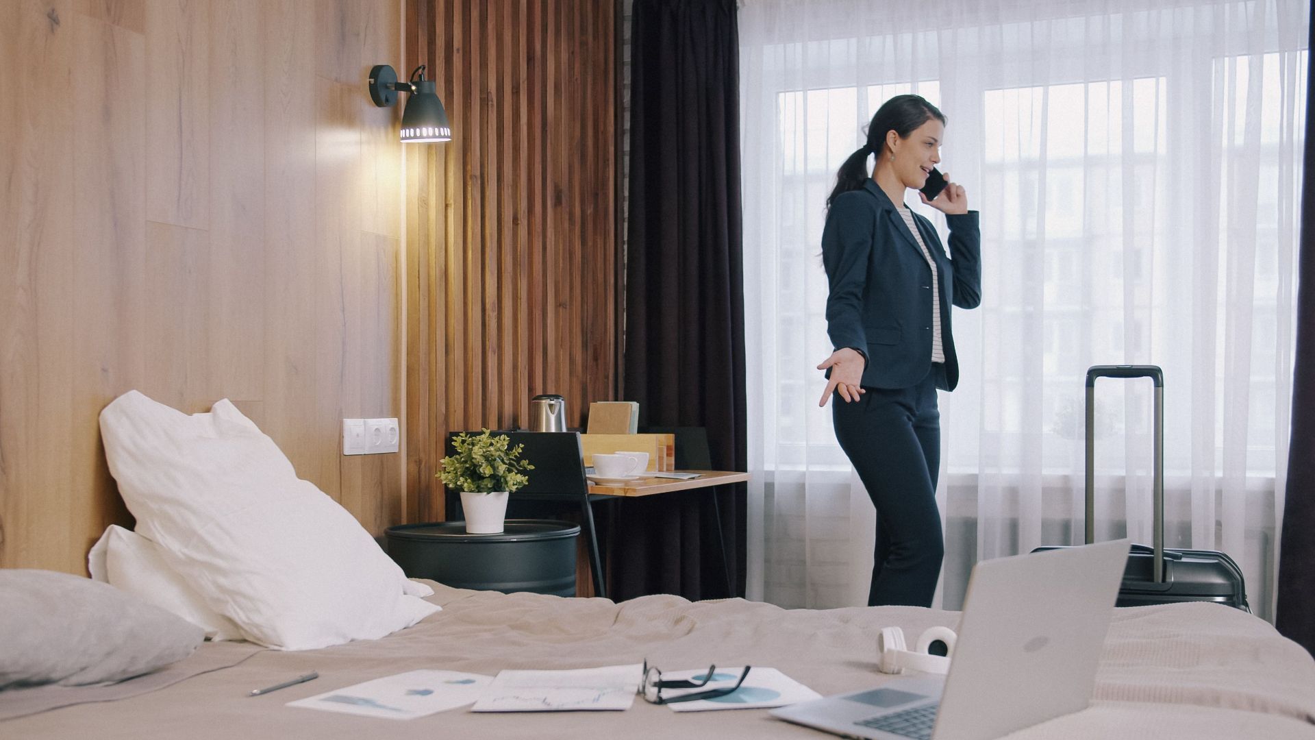 Professional woman multitasking in a hotel room, using a phone and laptop while traveling.