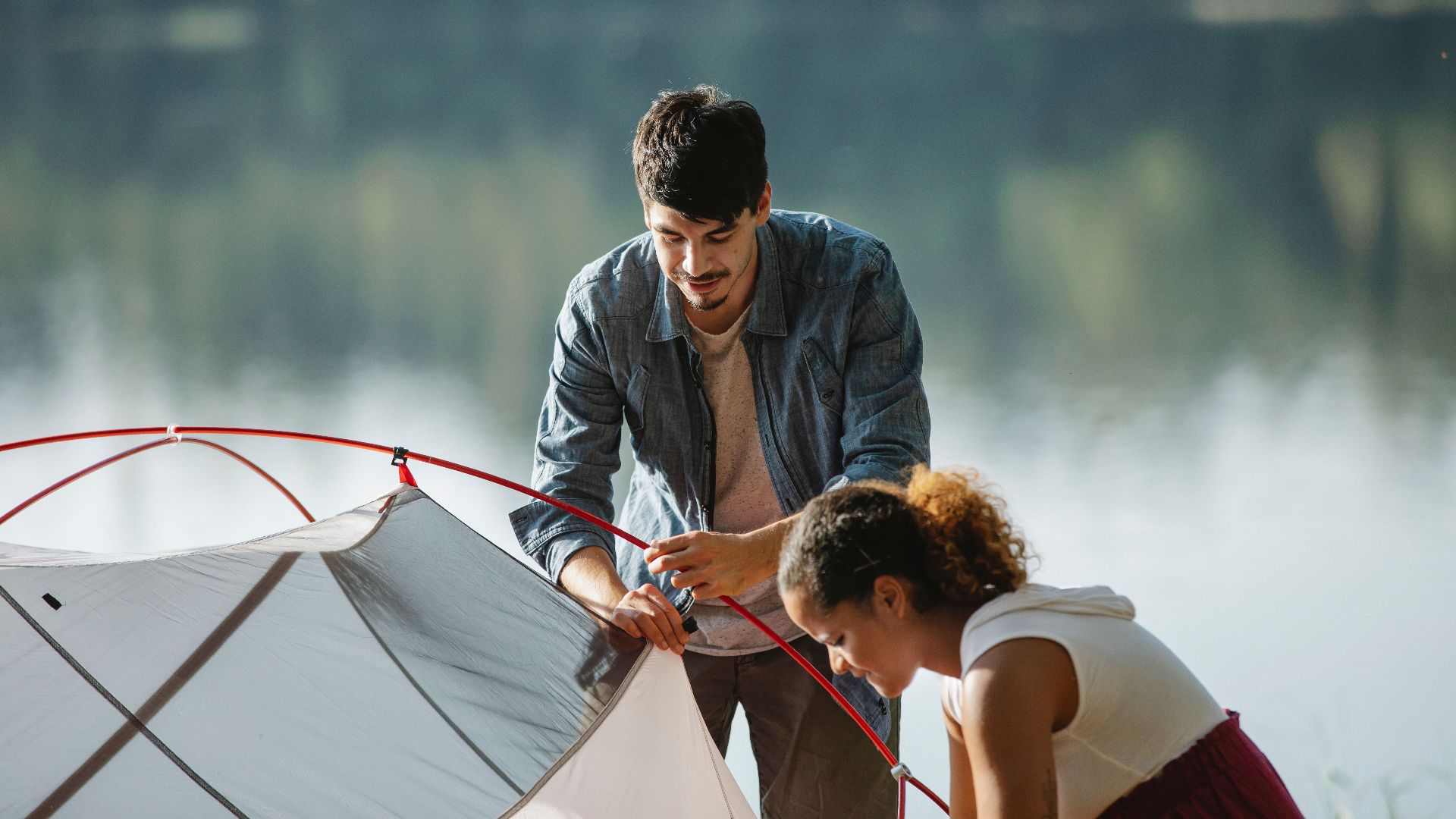 Concentrated young multiracial couple of travelers in casual wear busy with putting up near river