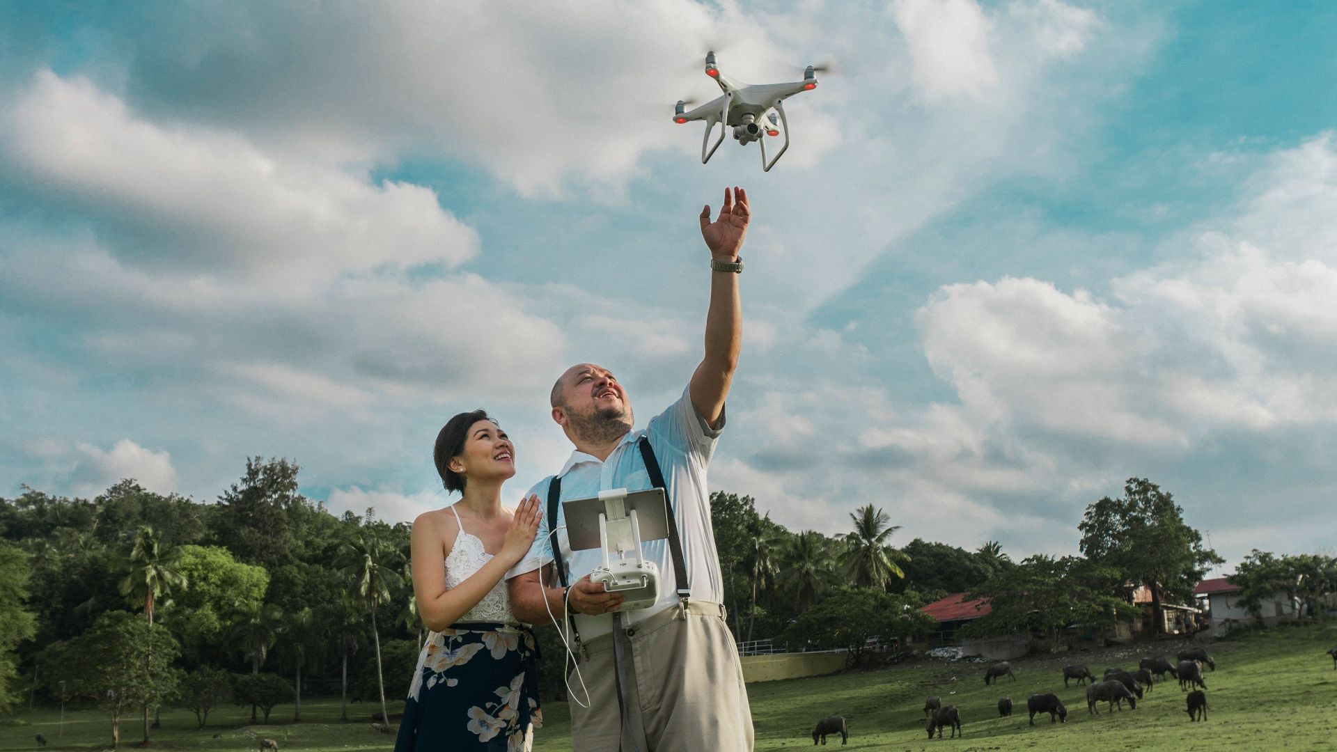 A couple enjoying their time flying a drone in a sunny park with clear skies and lush greenery.
