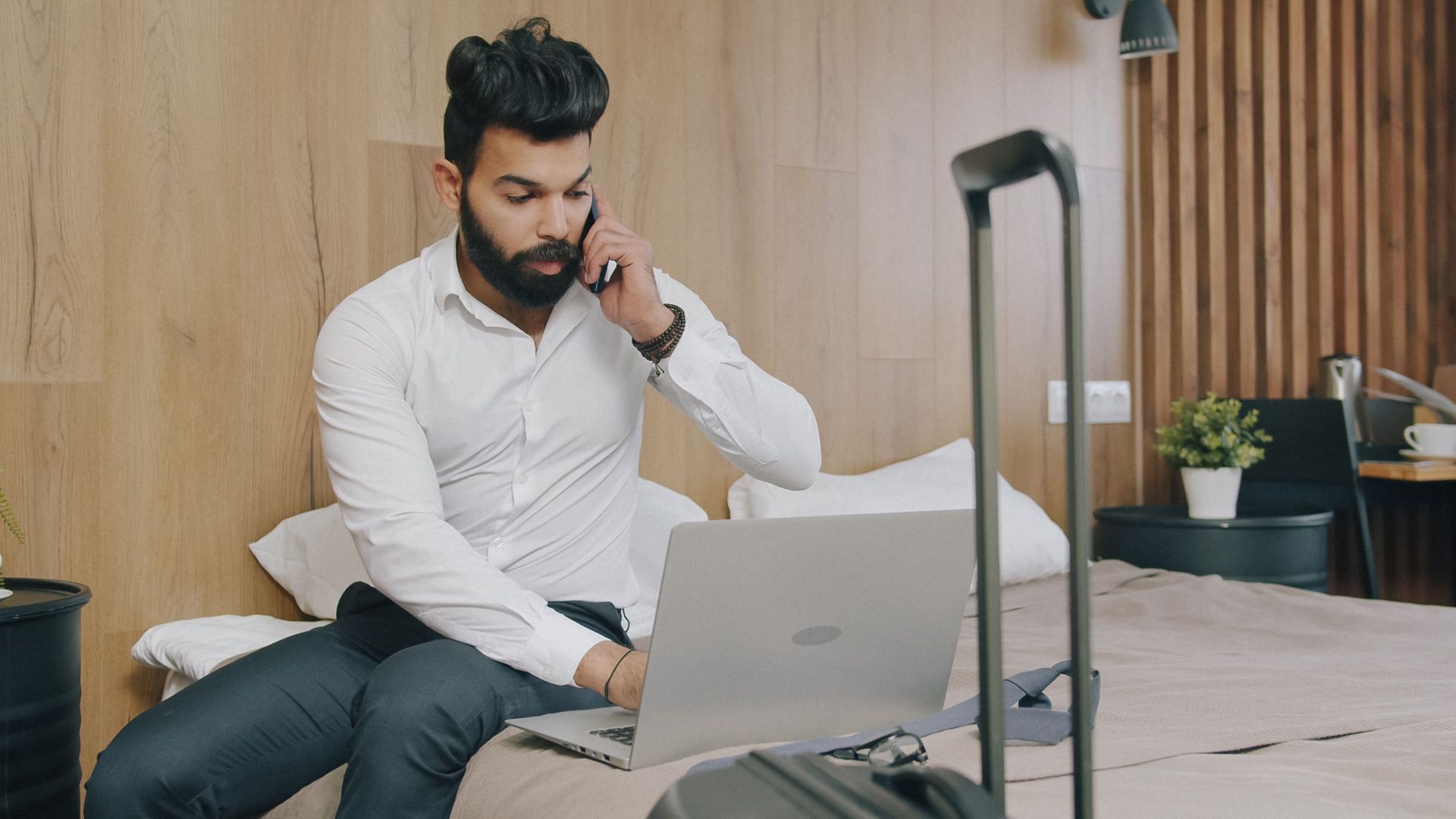 Businessman on laptop and phone in hotel room with luggage, working remotely.