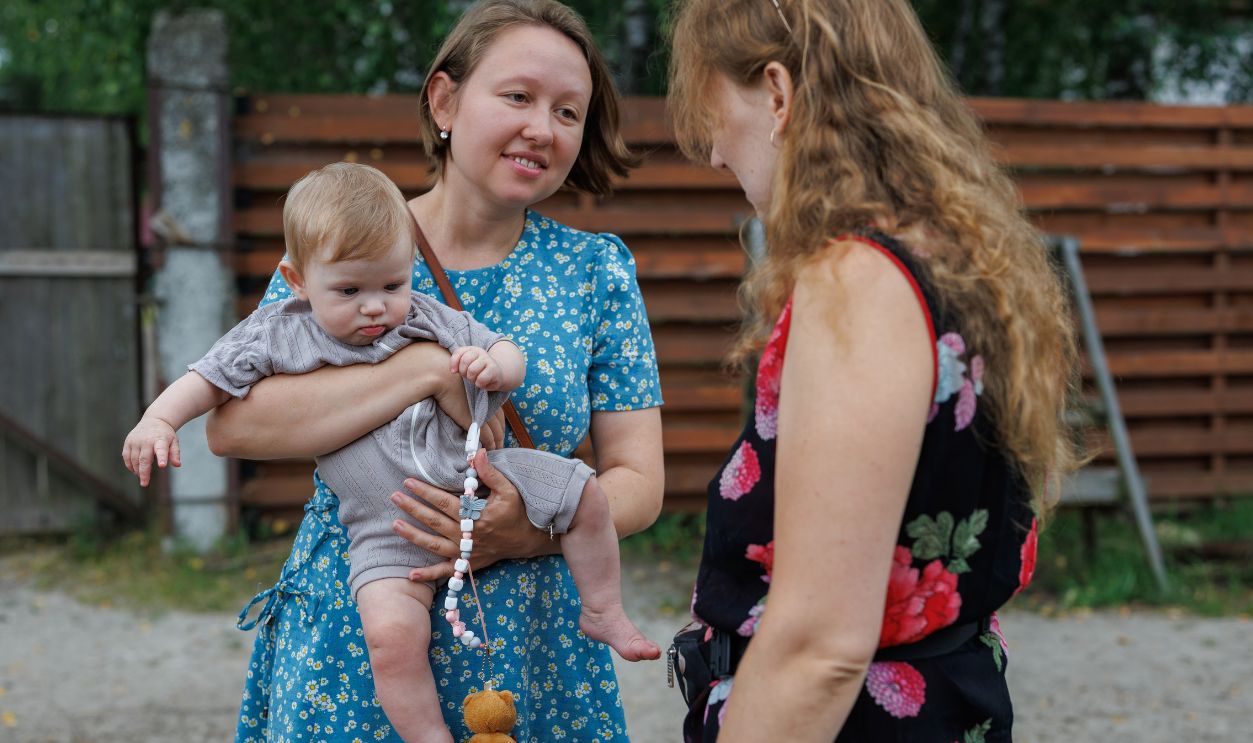 A woman in a blue dress holding a baby while talking to another woman outdoors near a wooden fence.