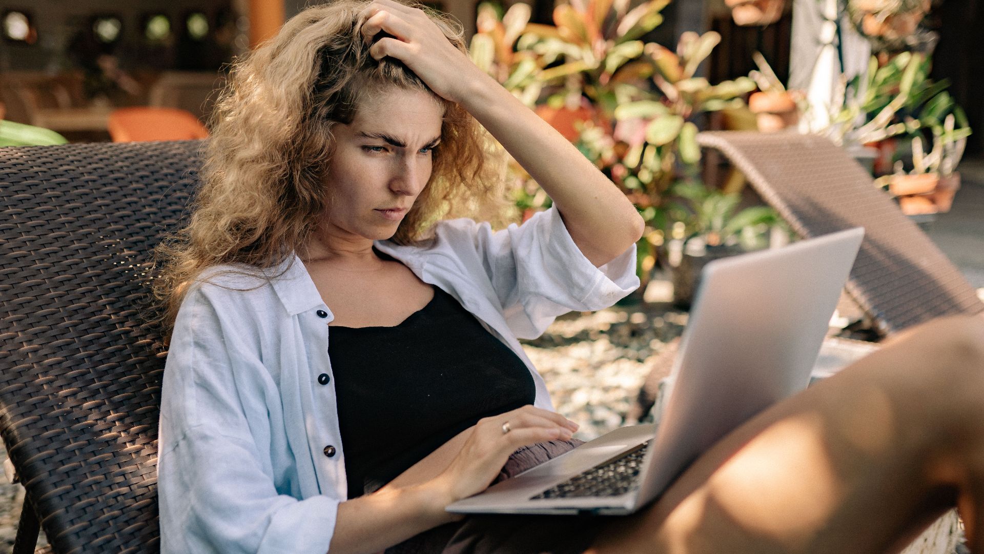 Focused woman with curly hair working on a laptop outdoors in a sunlounger, embracing a digital nomad lifestyle.
