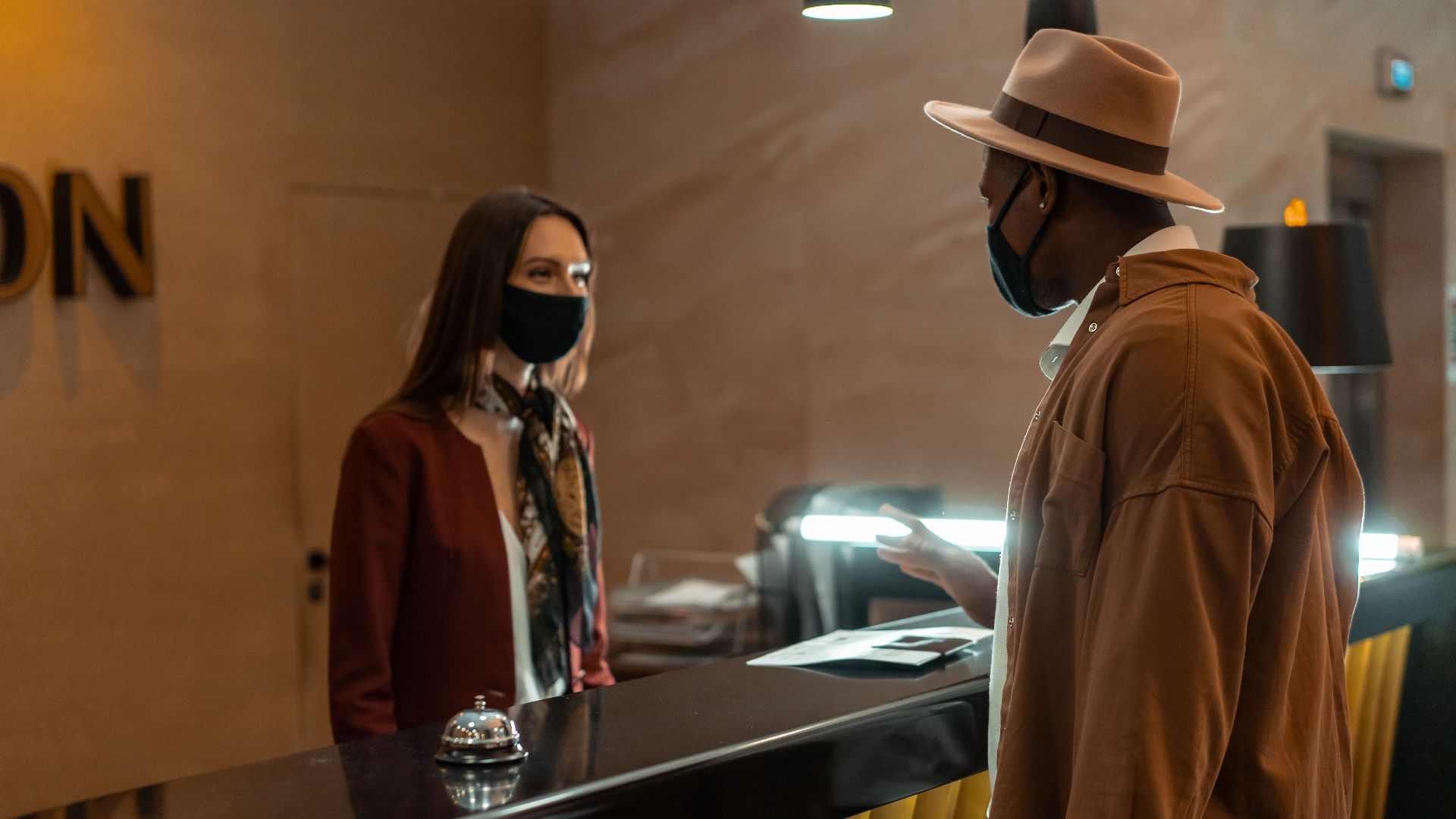 A man at a hotel reception talking to a staff member. Both wear face masks.