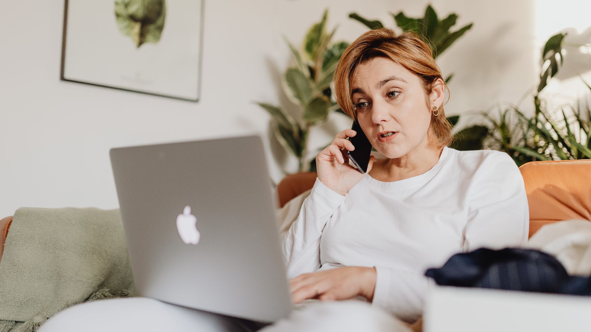 Adult woman engaged in multitasking at home using a laptop and phone, surrounded by plants.