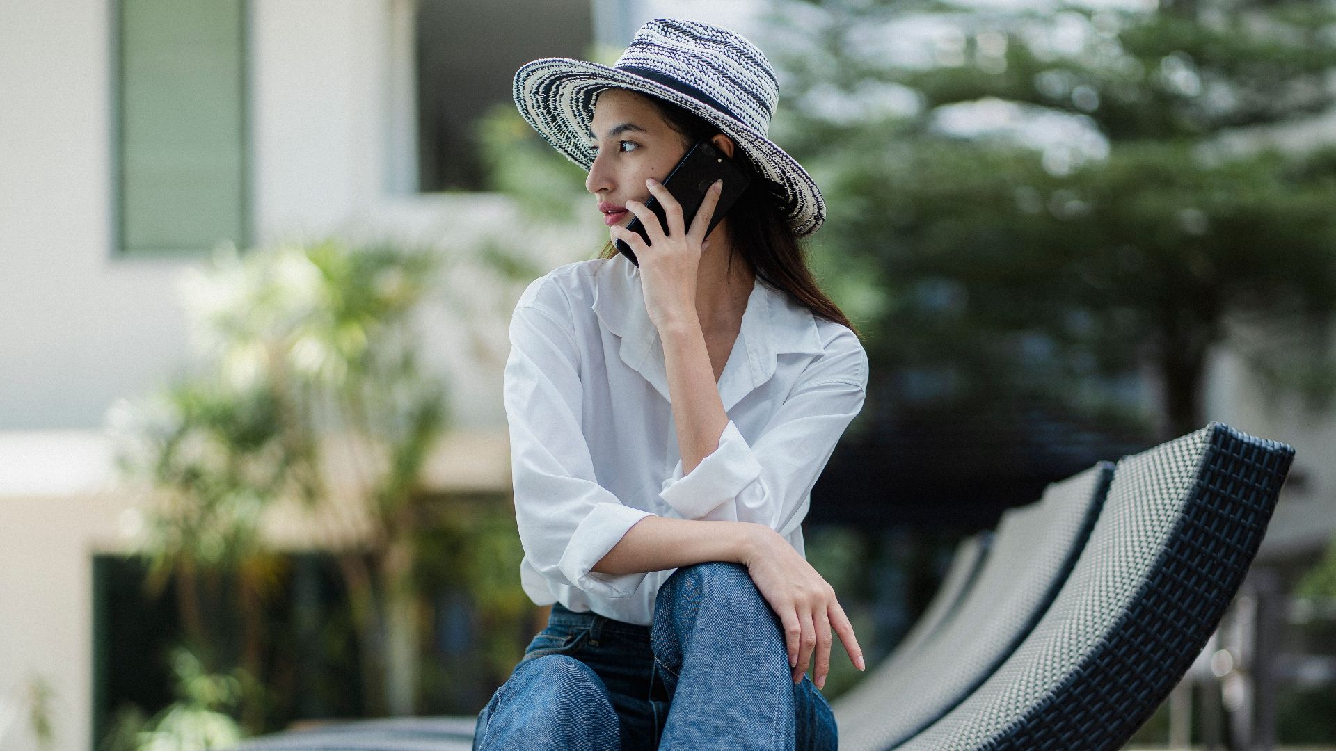 Full body of young stylish ethnic female in high heeled shoes and hat sitting on sunbed and having phone conversation in hotel garden