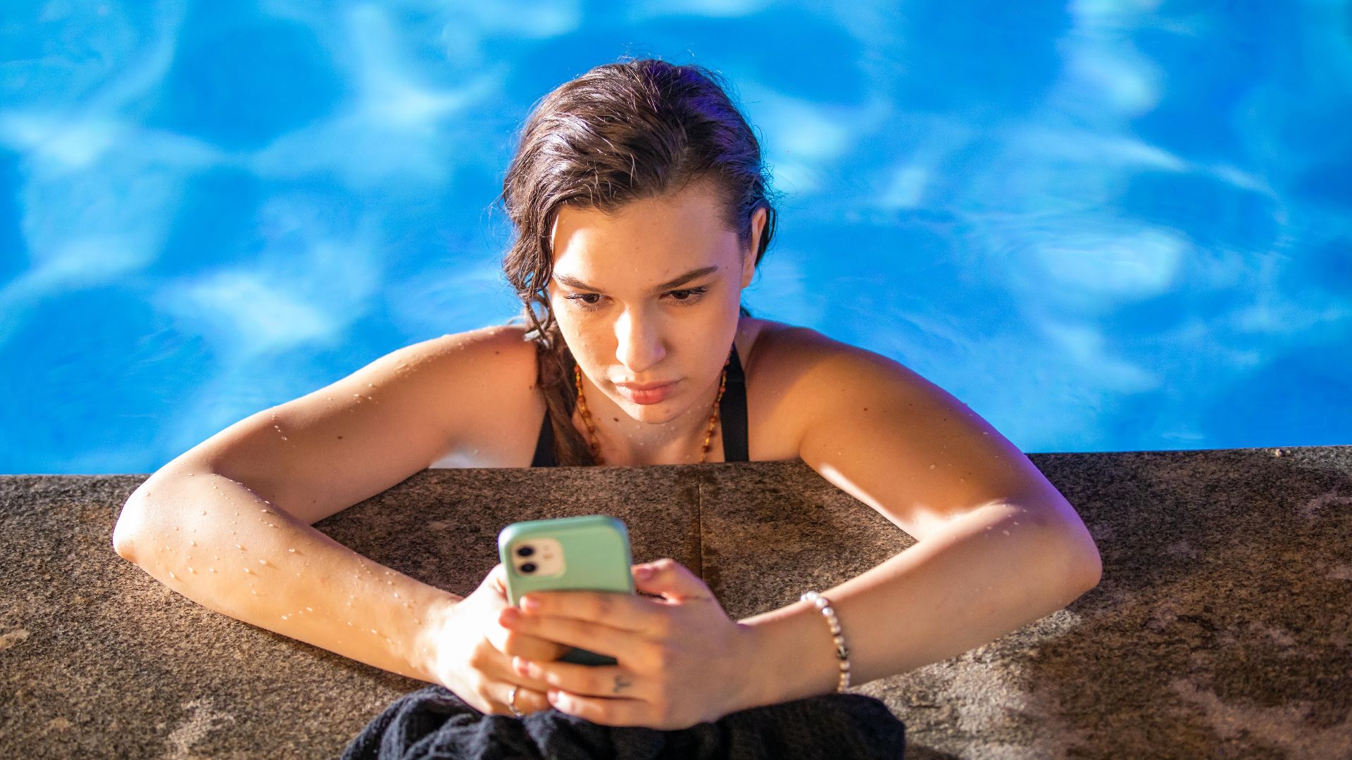 A young woman in a pool checks her smartphone under the bright sun.