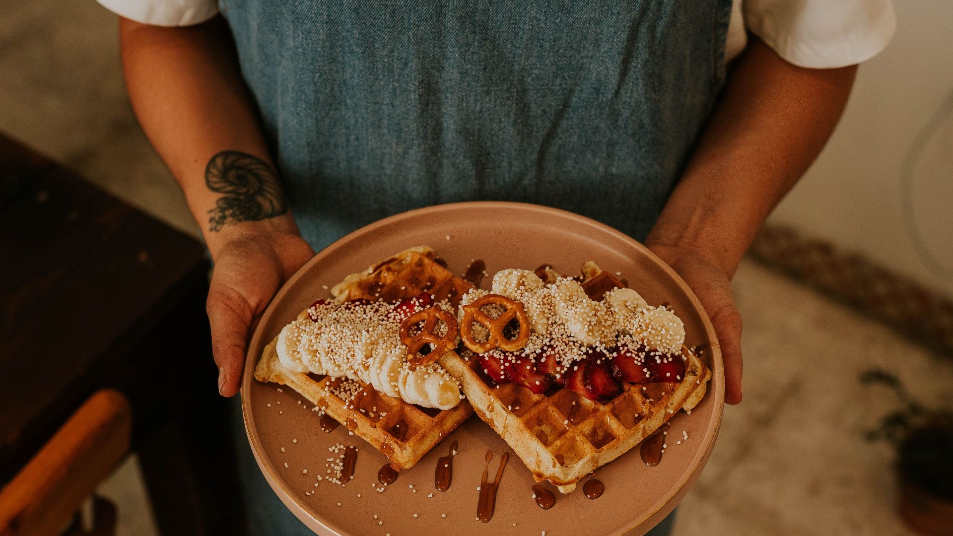 Close-up of a person holding a plate of waffles topped with cream, berries, and pretzels in a cozy kitchen setting.