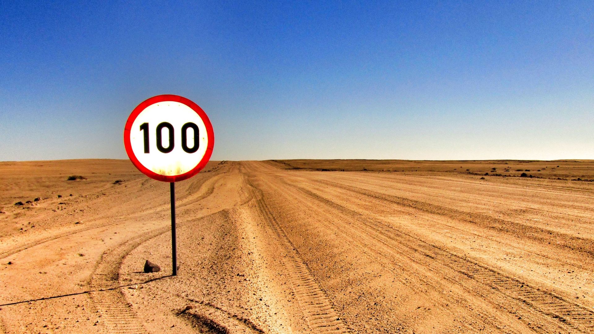 Open desert road with a speed limit sign showing 100 under clear sky.