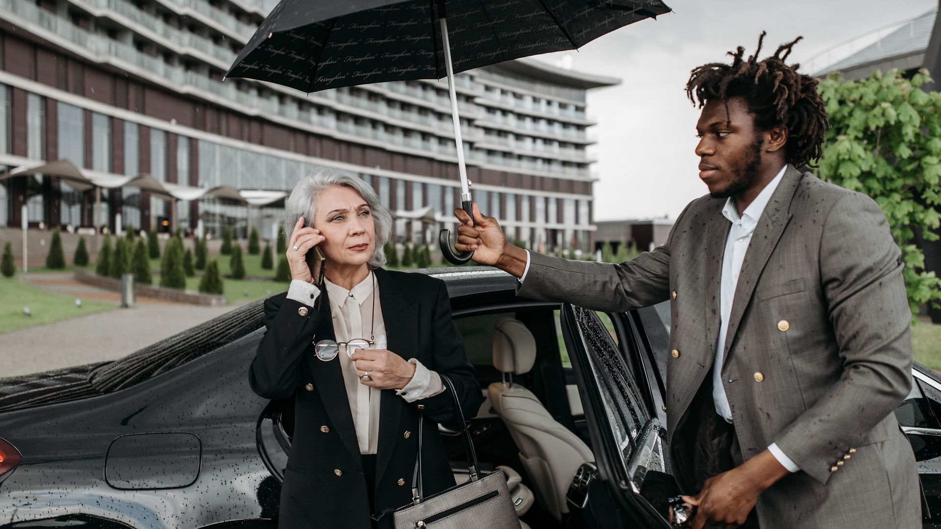 Two professionals, one holding an umbrella for the other, near a building on a rainy day.