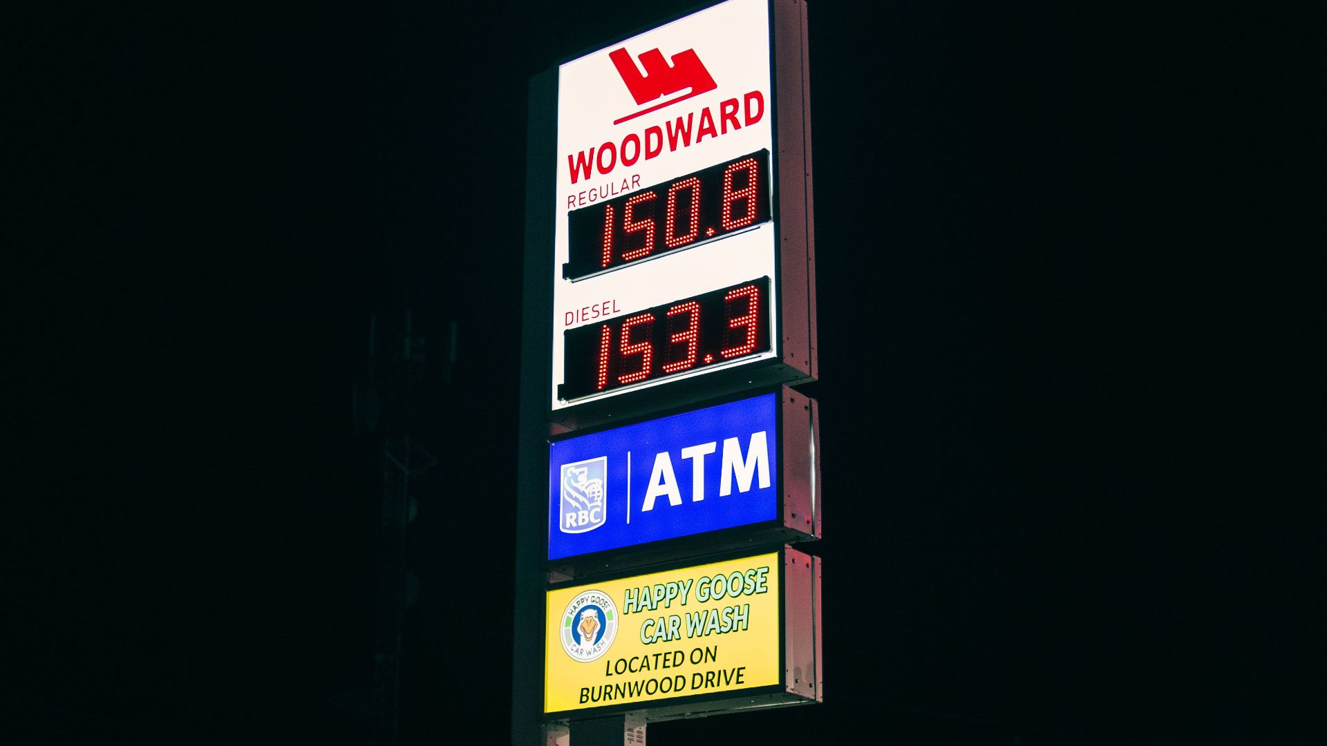 Gas station sign illuminated at night displaying fuel prices and ATM availability.