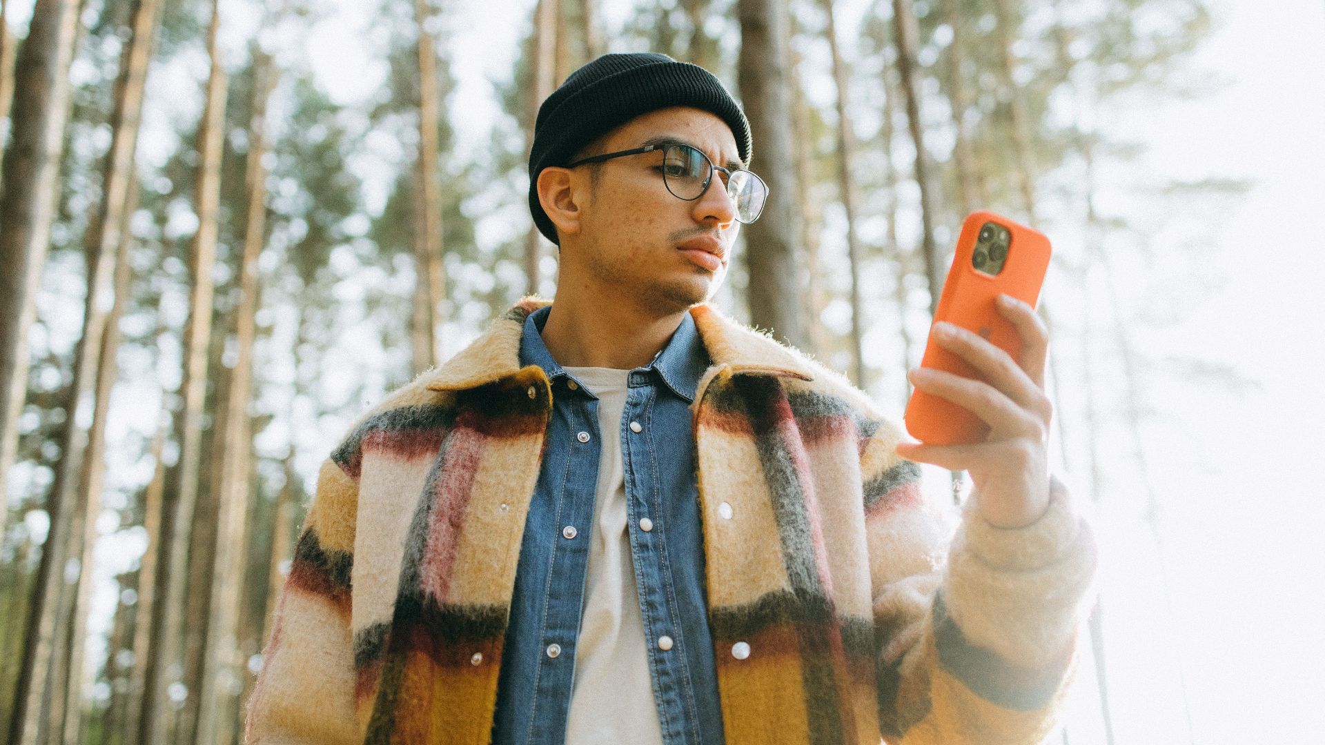 A trendy man in a beanie and plaid coat checks his smartphone amidst tall forest trees.