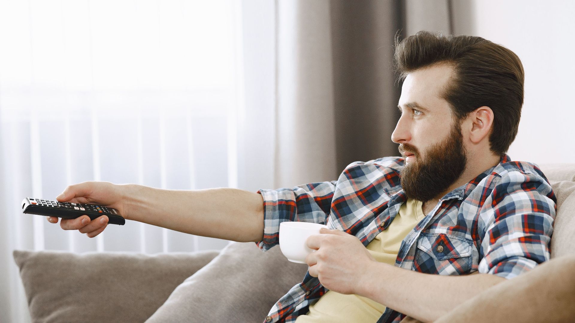 Bearded man in casual wear relaxing on a sofa, enjoying coffee while watching TV indoors.