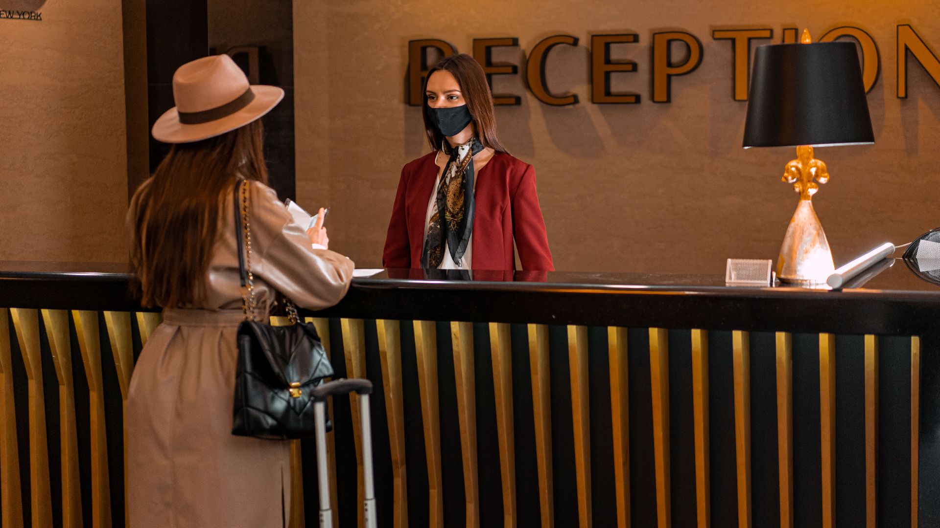 A woman speaks to a receptionist wearing a mask at a hotel reception.