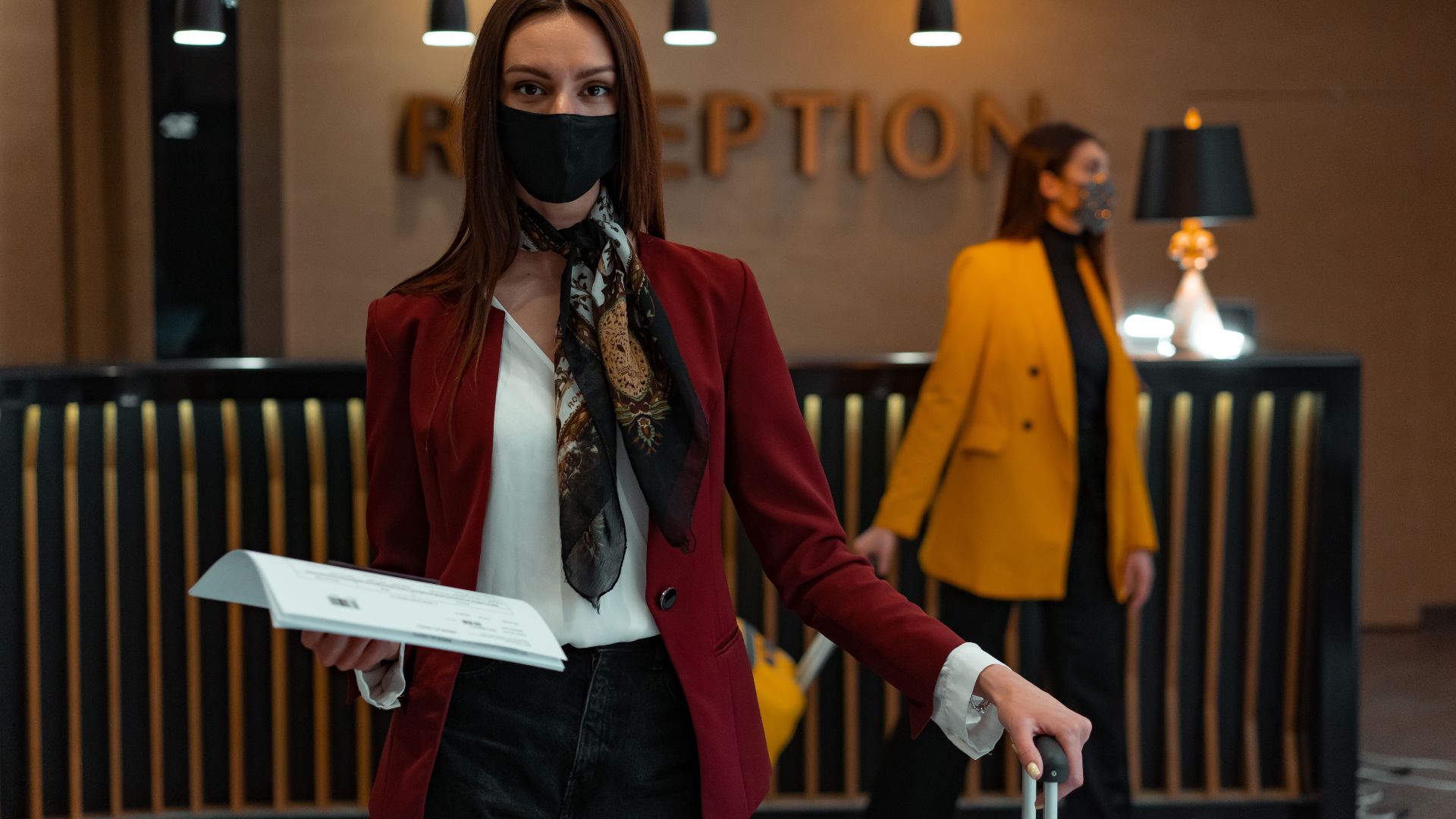 A woman in a mask stands at a hotel reception desk holding paper, welcoming service