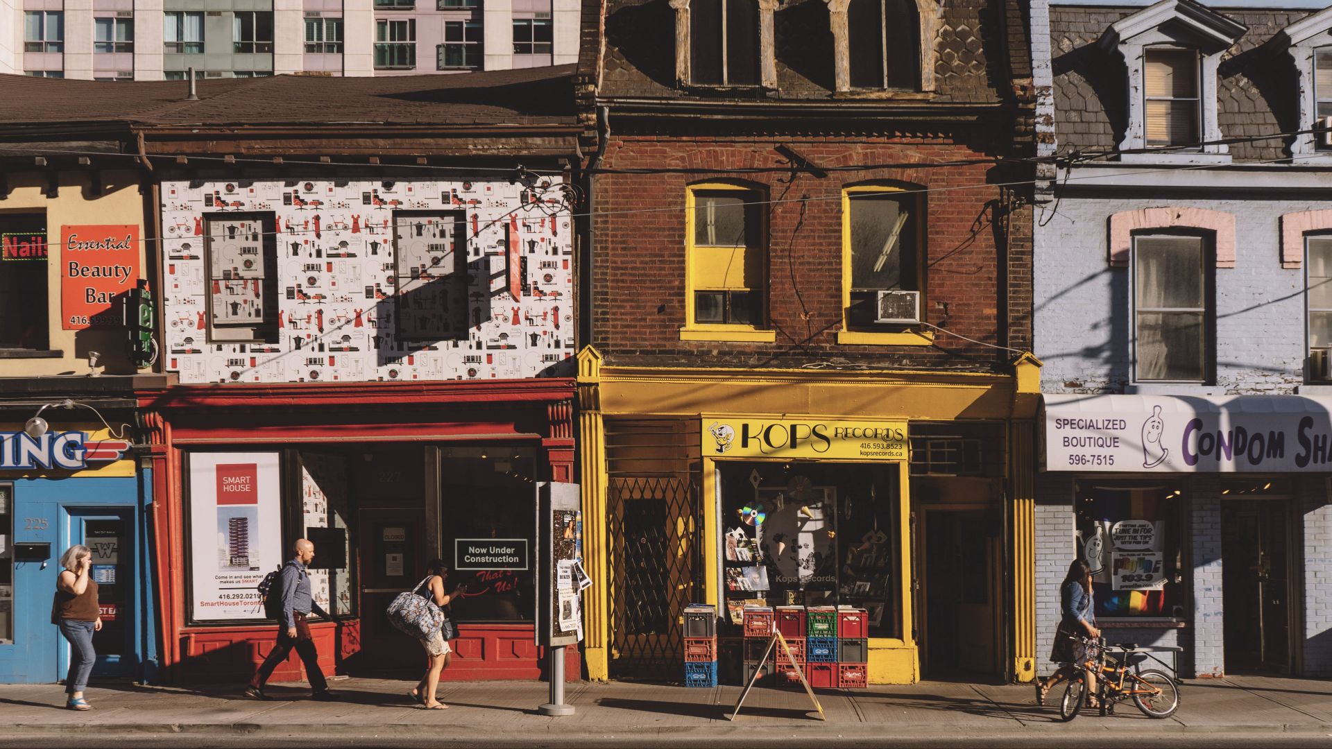 Colorful street scene with people walking past unique storefronts under sunlight.
