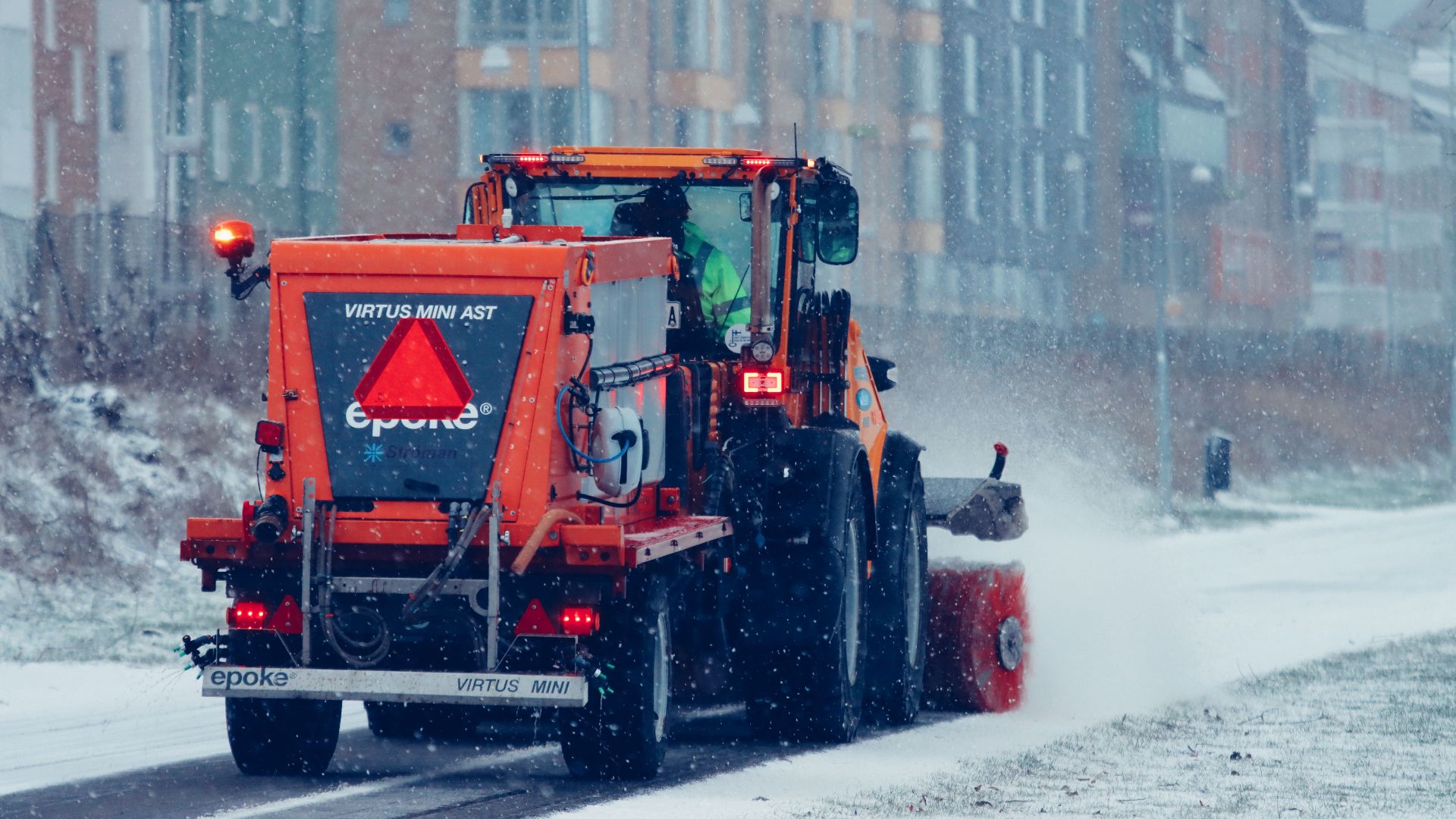 Orange snowplow in action clearing a snow-covered street during a winter snowfall.