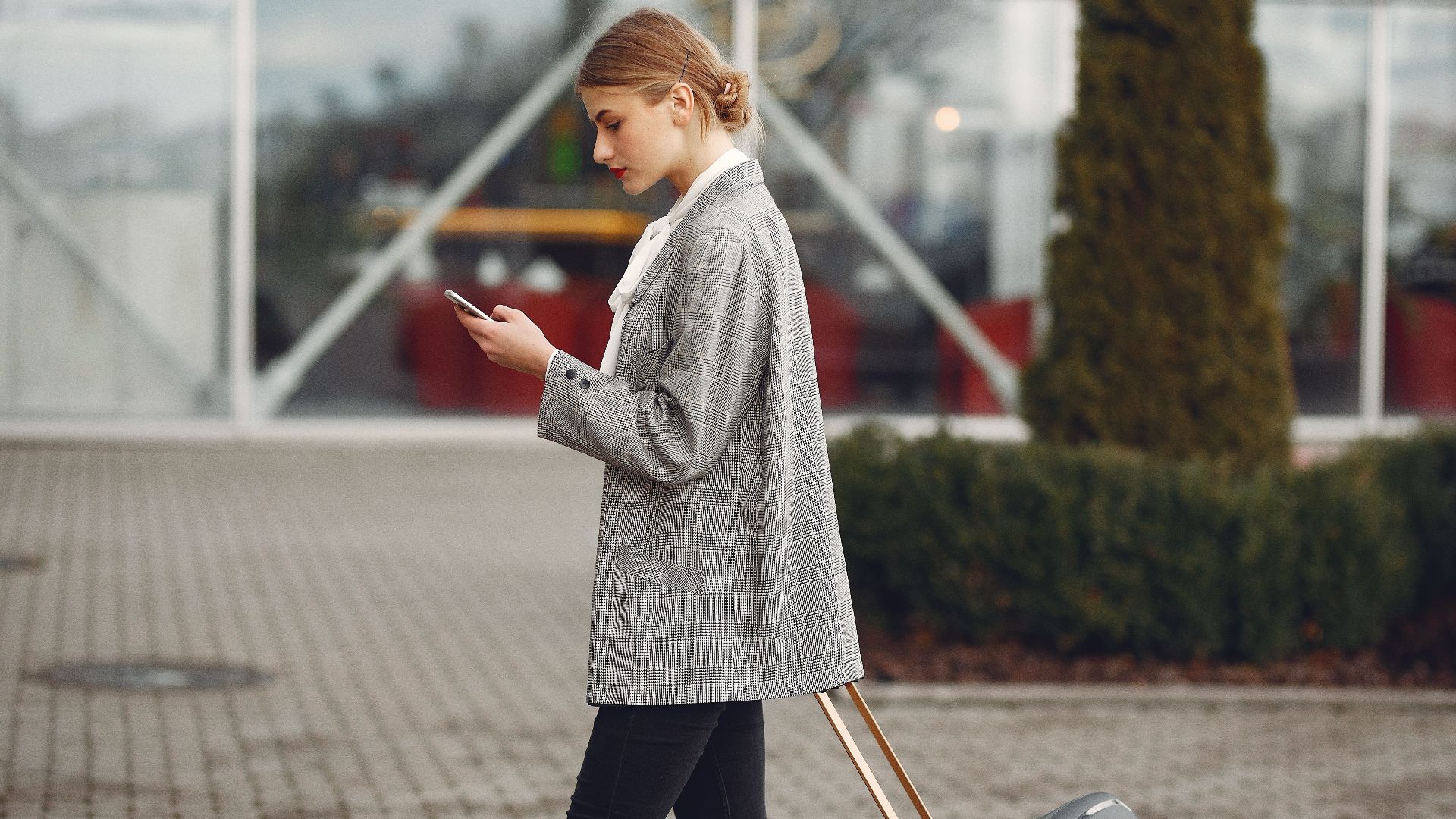 Stylish woman walking with suitcase while using smartphone outside a modern building.