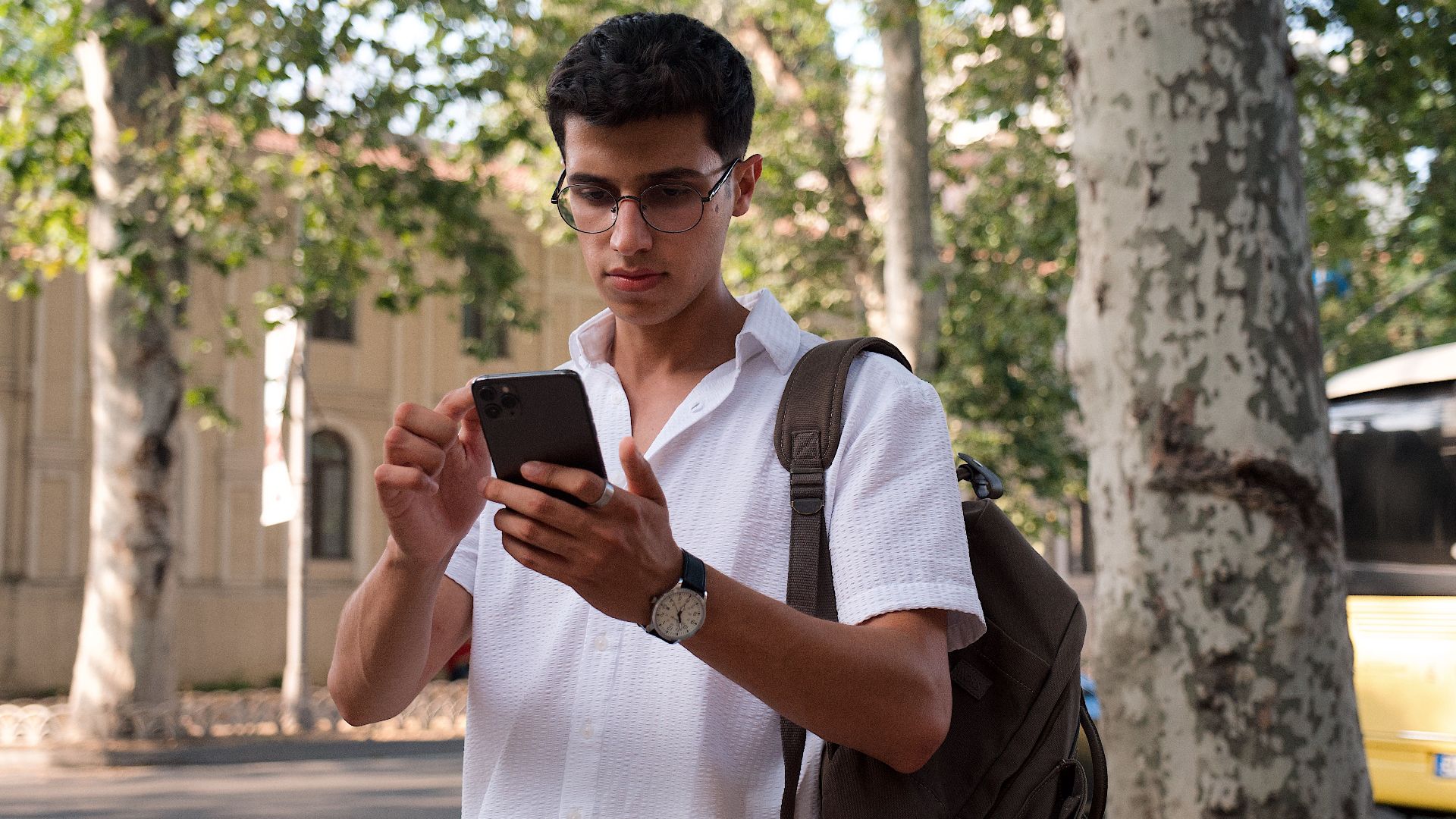 Young man using smartphone on a city street, wearing eyeglasses and a backpack.