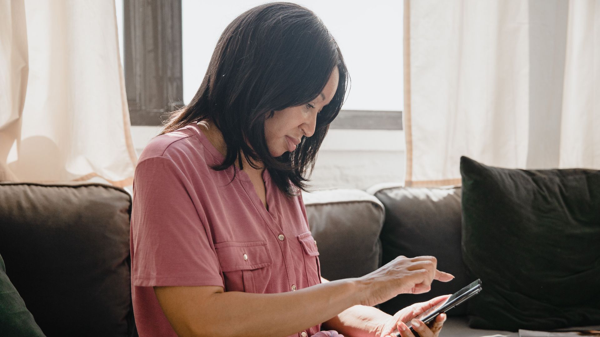 Woman in pink shirt using a smartphone while sitting on a couch, side view.