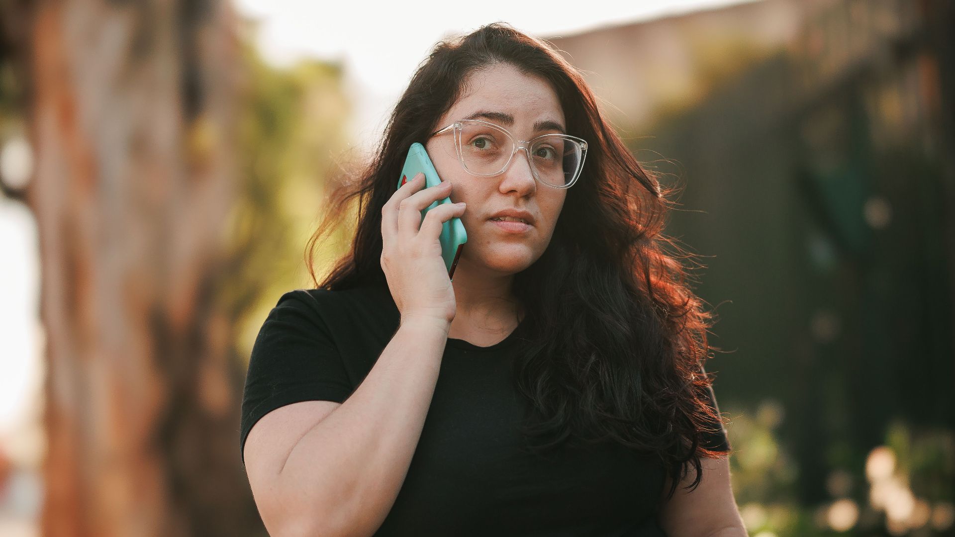 Woman with eyeglasses talks on smartphone in sunny park setting.