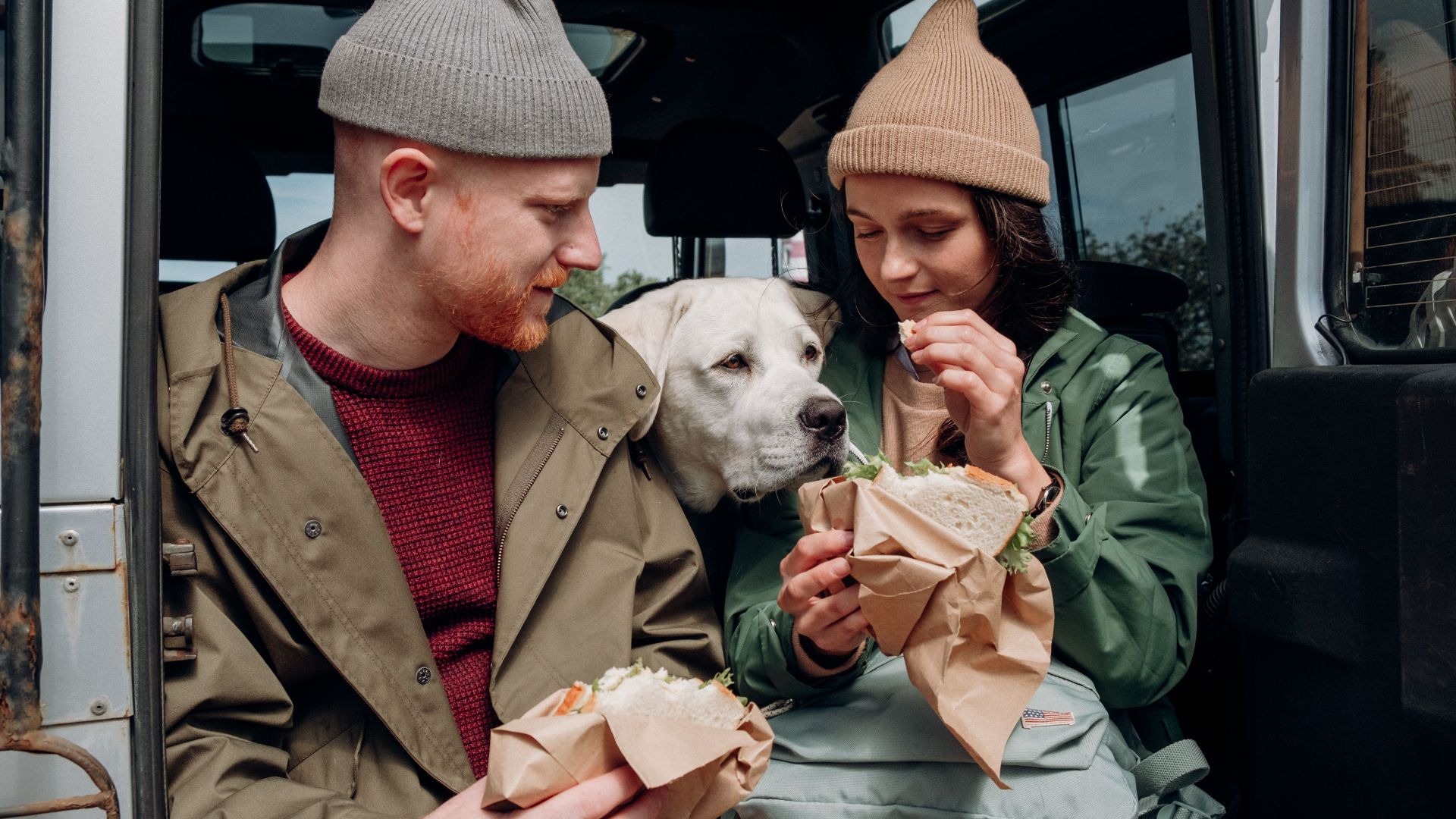 A couple enjoys sandwiches with their dog during an outdoor adventure.