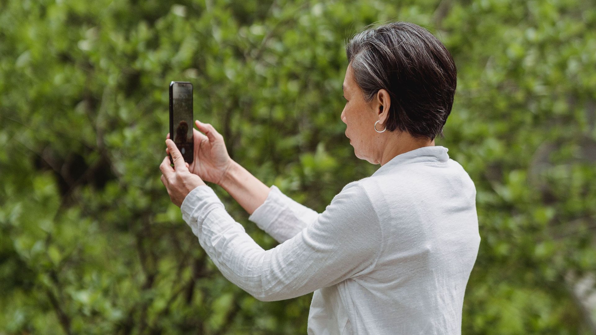 A woman in a white shirt takes a photo with her smartphone in a lush green park.