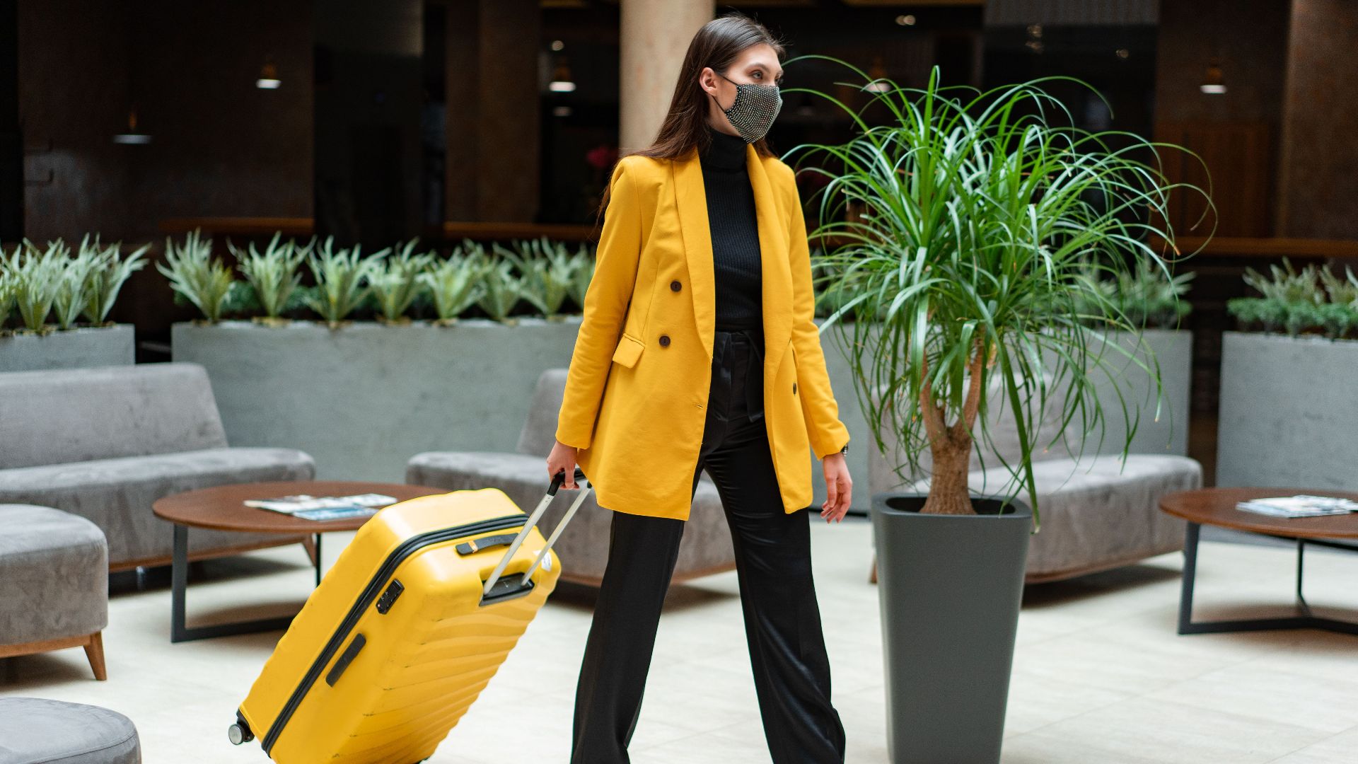 Fashionable woman in yellow blazer and face mask walking with suitcase indoors.