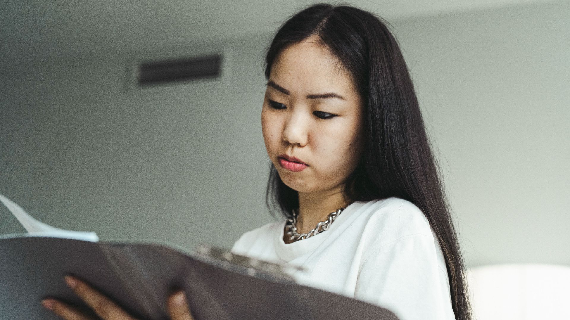 Asian woman in casual attire examining paperwork with concentration indoors.