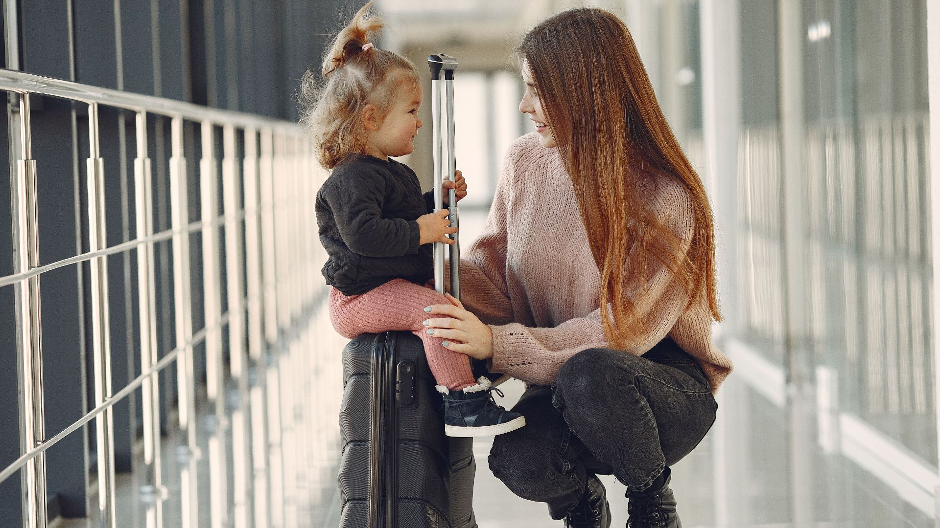 Full length of positive content woman and cute stylish blond little girl sitting on black suitcase in airport corridor while waiting for flight