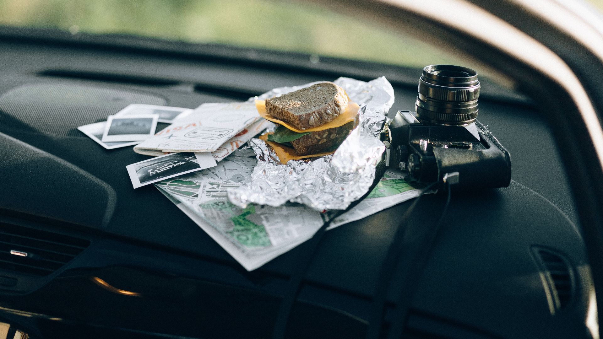 A camera and a sandwich wrapped in foil placed on a car dashboard with maps and photos.