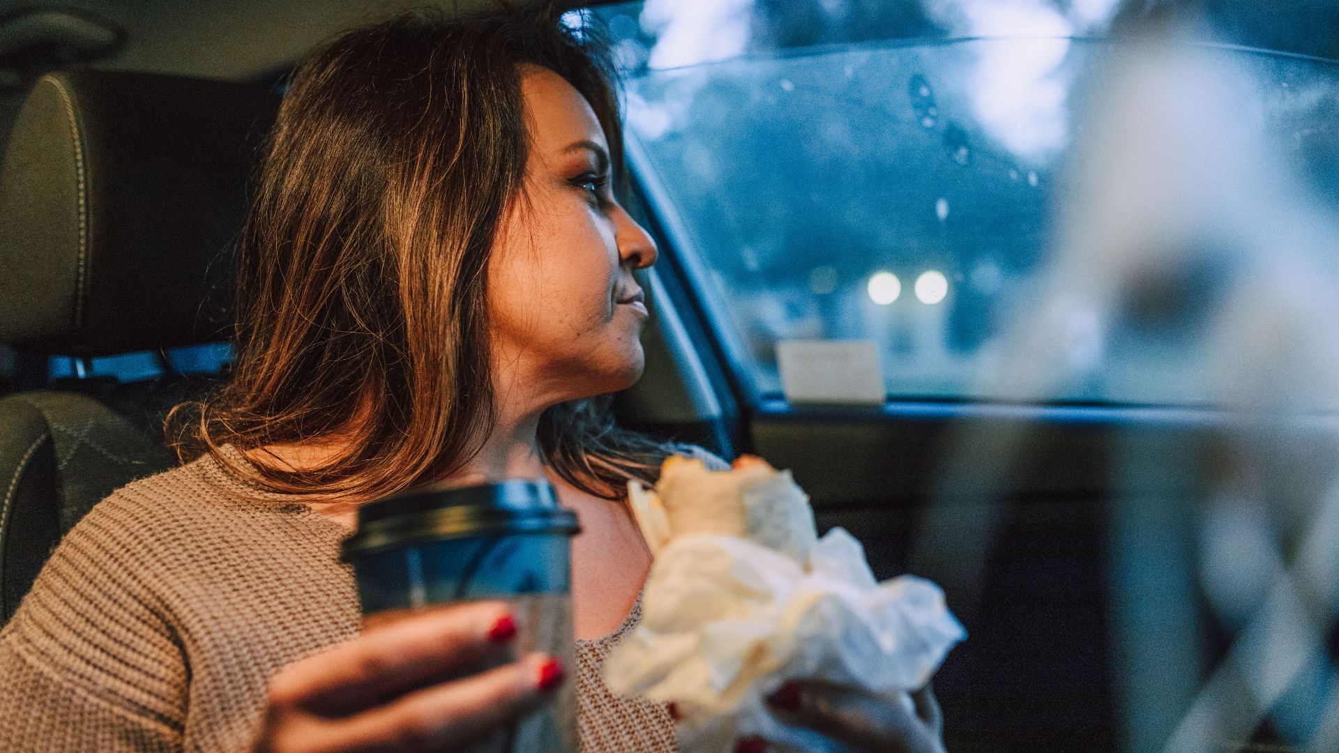 A woman sits in her car with a coffee and a sandwich, reflecting on her day.