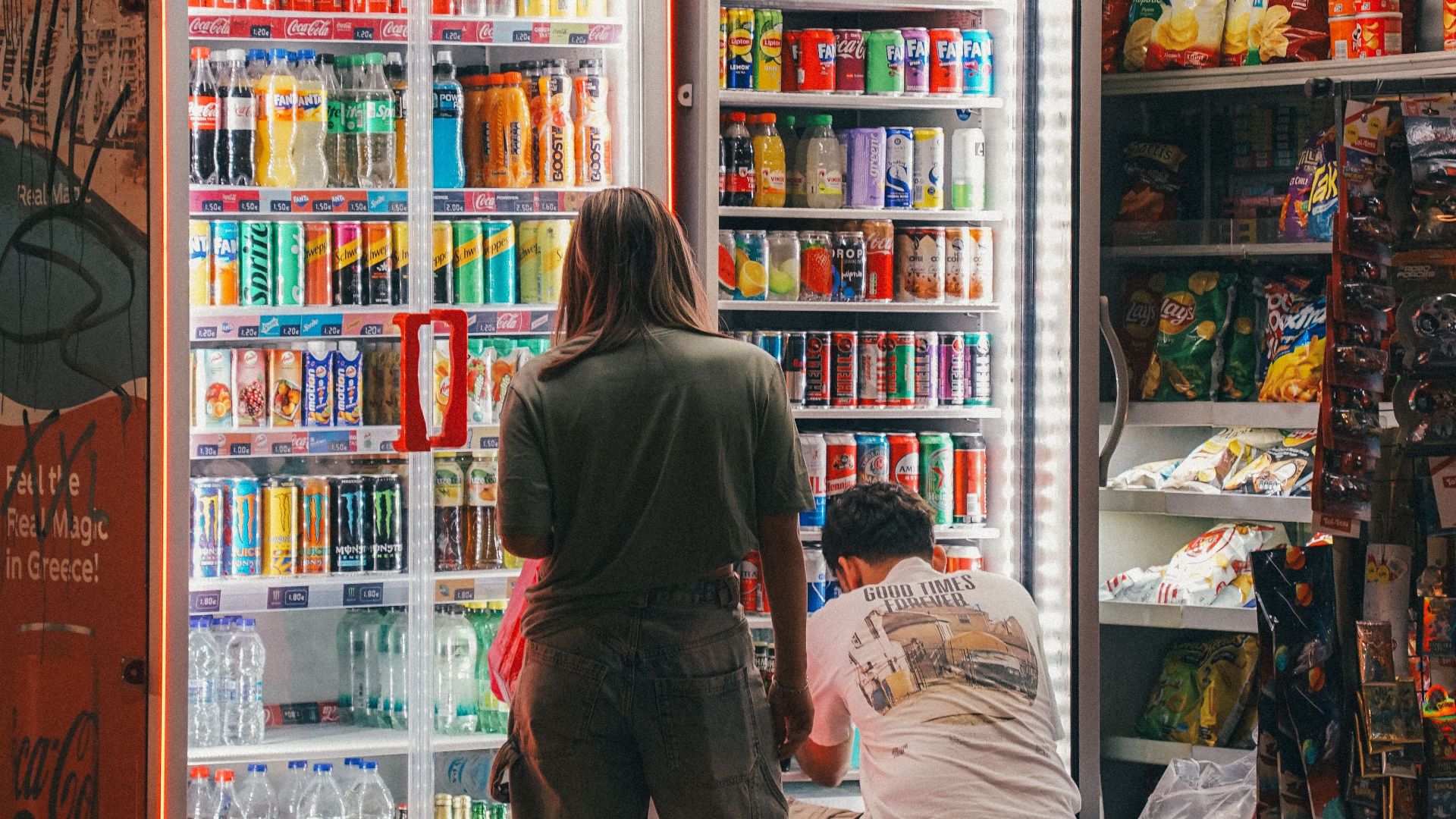 People selecting drinks at a convenience store aisle illuminated by vending machines.