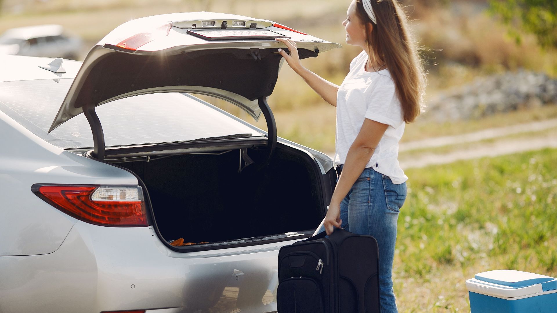 Side view of cheerful female traveler in headband and casual clothes putting luggage in open trunk of modern car while spending summer weekend in countryside