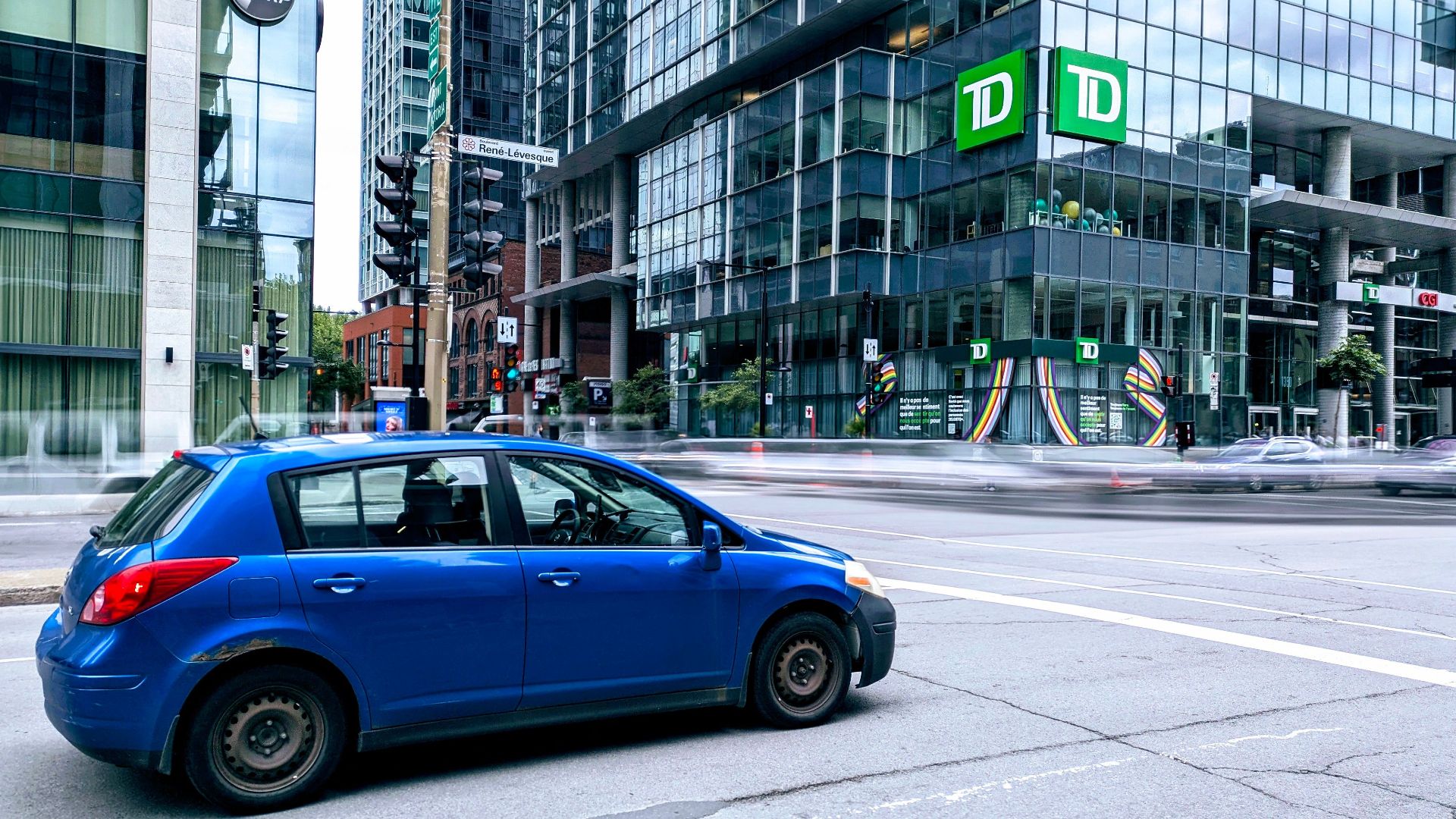 Blue car parked on city street with towering glass skyscrapers in the background.