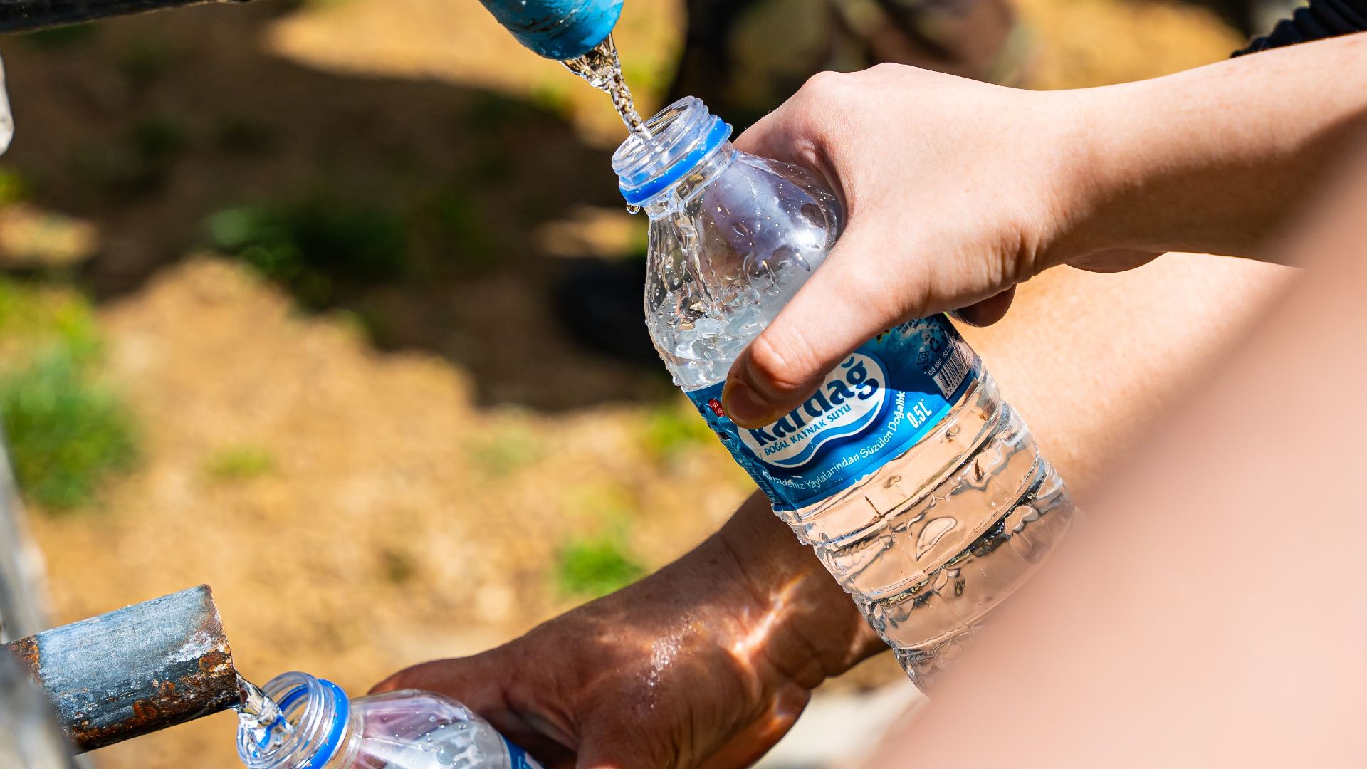People filling water bottles from an outdoor tap in Düzköy, Trabzon, Türkiye, on a sunny day.