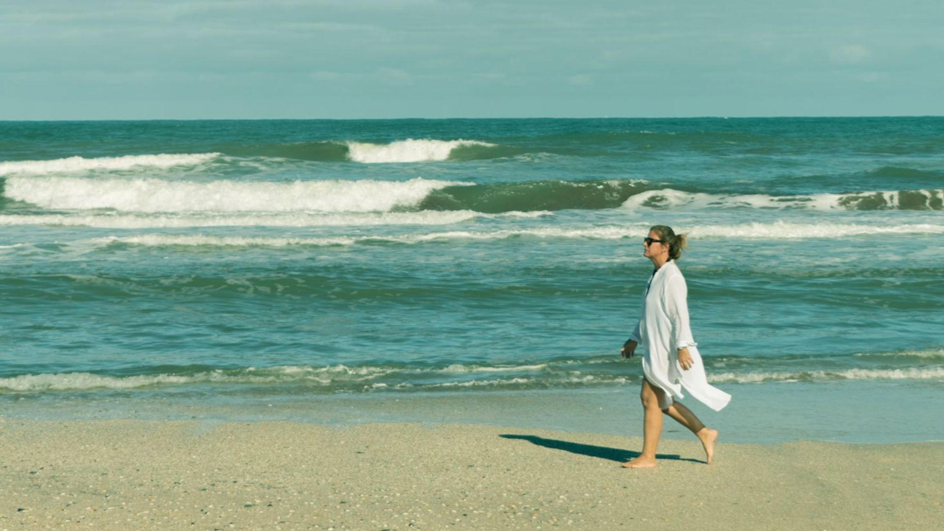 A woman in white walks barefoot along a serene beach under a clear blue sky.