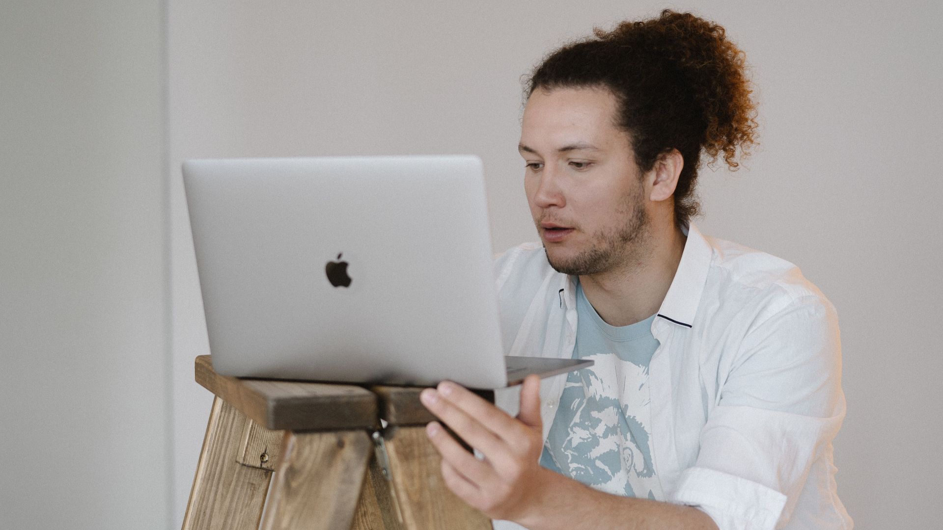 A man using a laptop while standing on a wooden ladder in a minimal indoor setting.