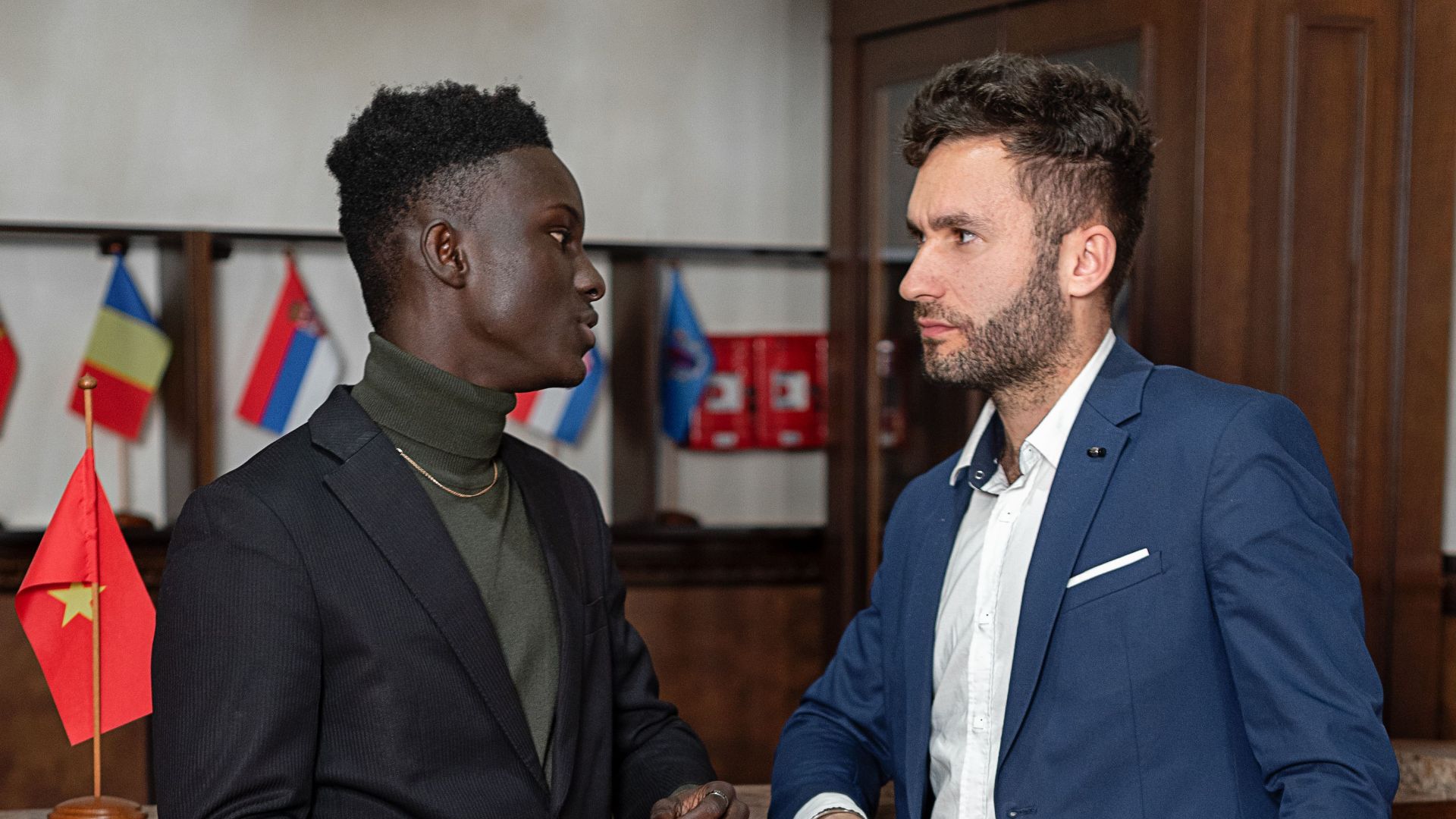 Two businessmen in suits having a conversation indoors, displaying flags in the background.
