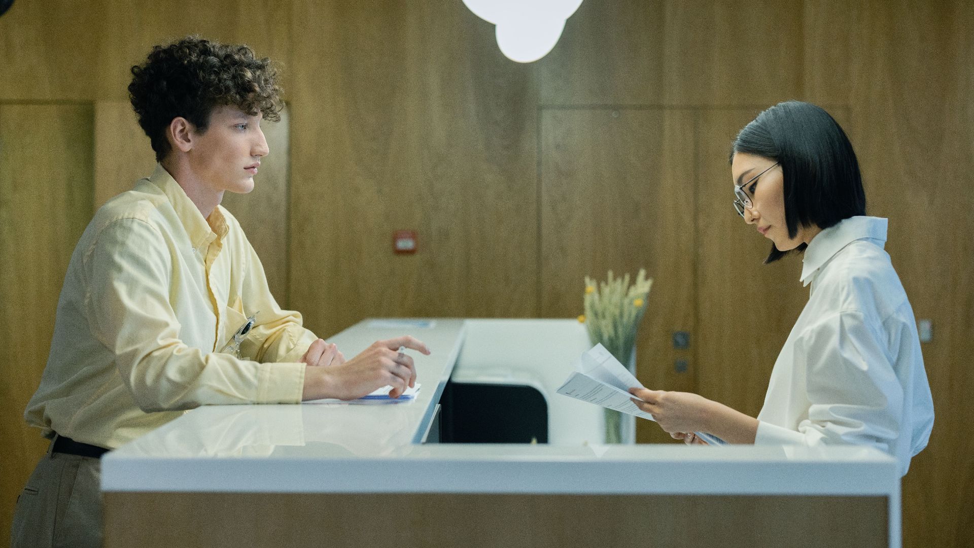 A young man and woman interacting at a modern office reception desk indoors.
