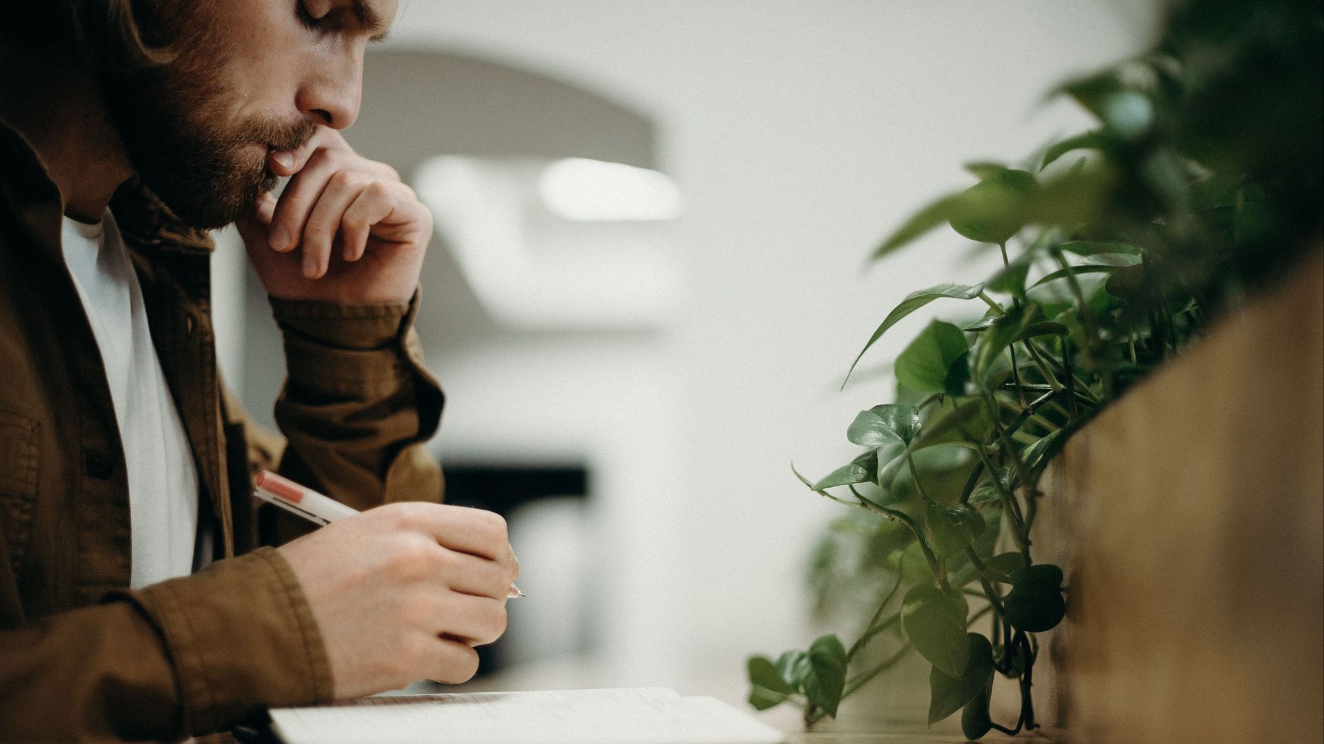 A man deeply concentrates while writing in an indoor office with plants.