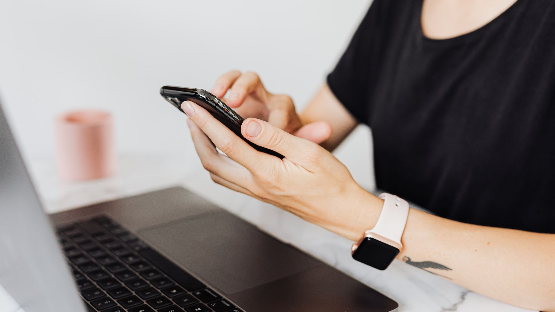 Close-up of a woman using a smartphone with a laptop on an office desk, showcasing technology in the workplace.