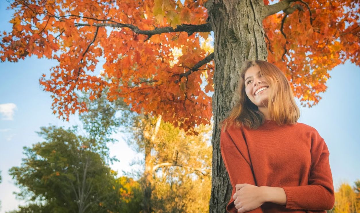 Woman in red sweater leaning against a tree with vibrant autumn foliage, smiling and enjoying the sunny day outdoors