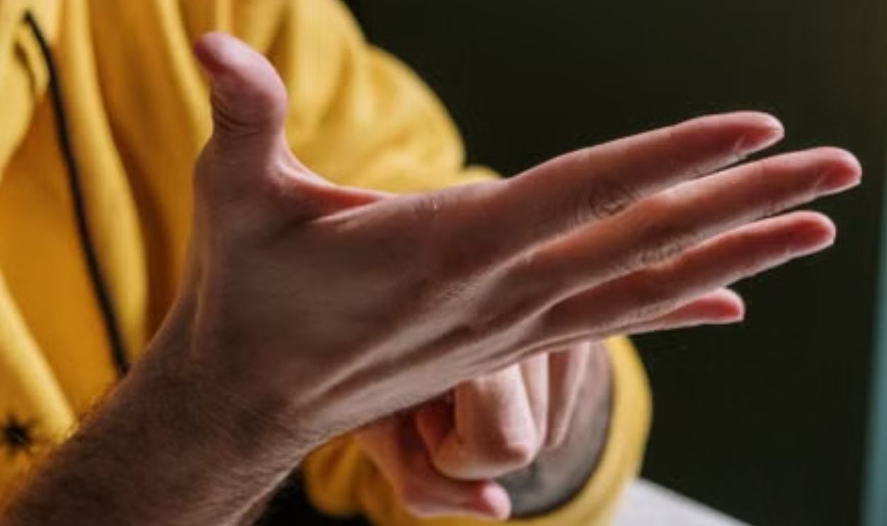 A man wearing a yellow hoodie uses sign language while sitting at a table indoors