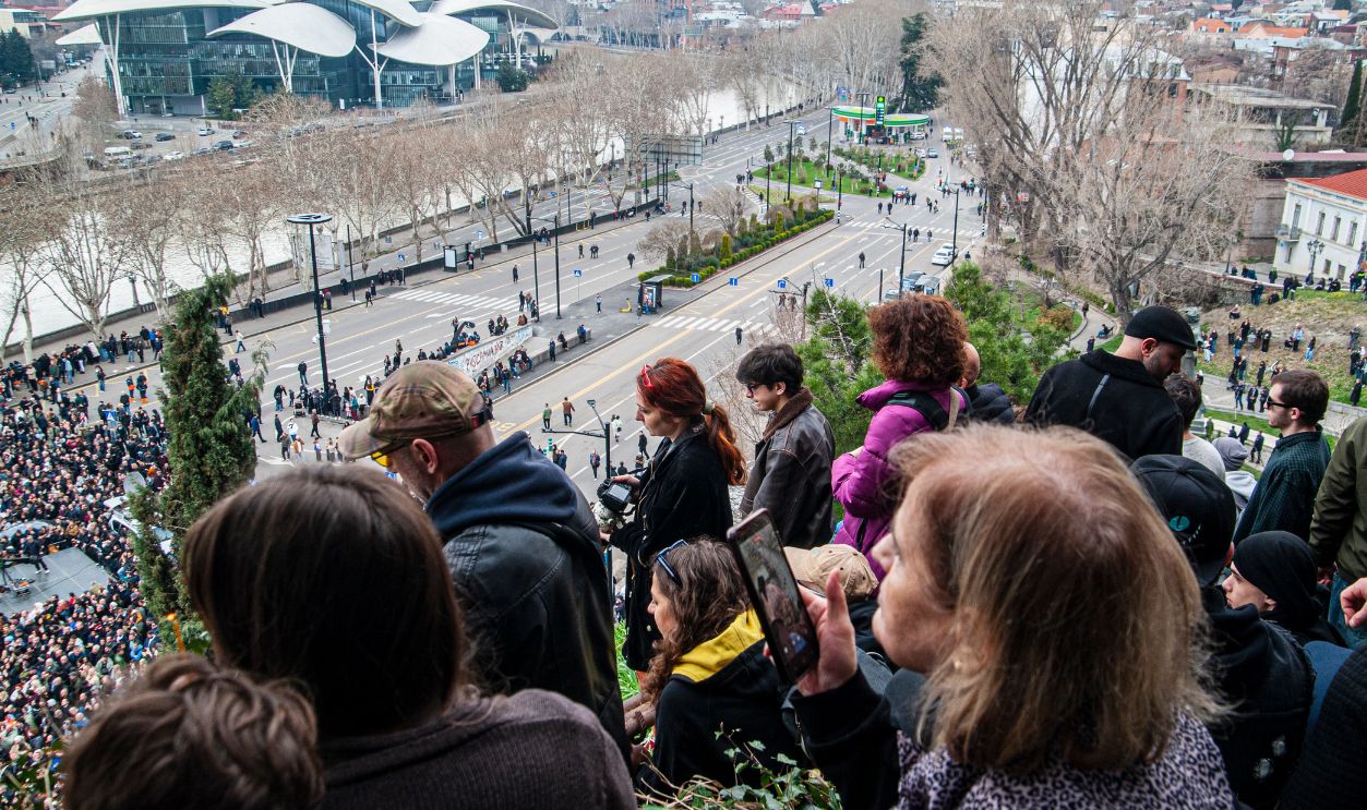 A large crowd gathers on an elevated area, overlooking a cityscape with modern architecture and a roadway