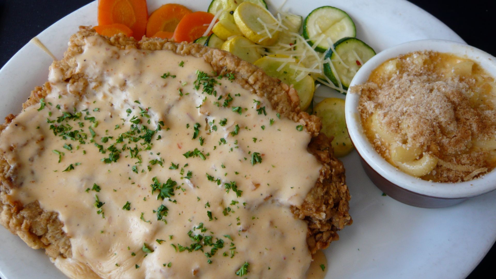 A white oval plate with chicken fried steak covered with chipotle cream gravy, macaroni and cheese, and vegetables (carrots, yellow squash, and zucchini) as served by the Moonshine Patio Bar & Grill at 303 Red River Street in Austin, Texas, USA.  Accordin